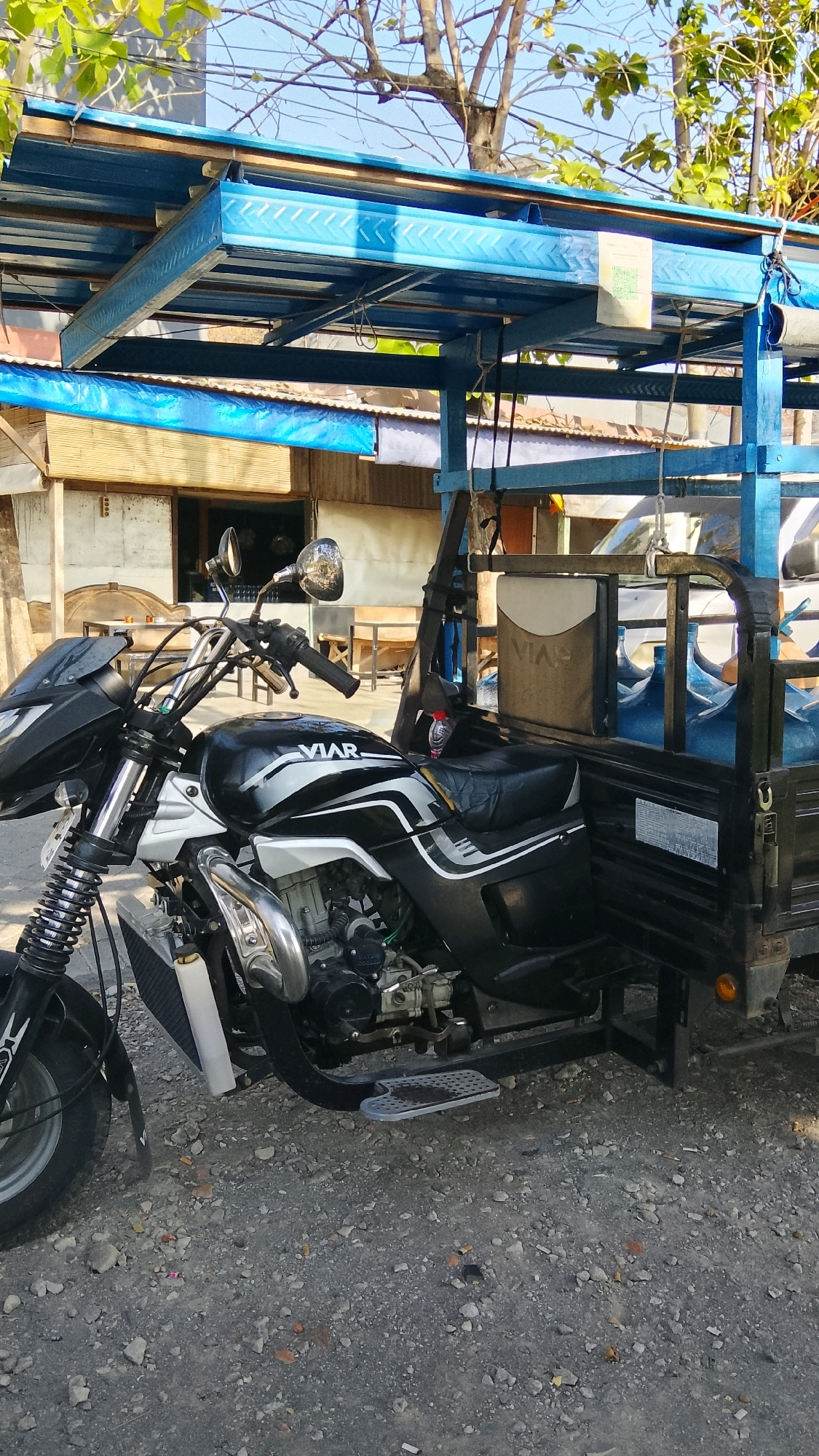 A side view of a black and white three-wheeled motorcycle, possibly a VIAR brand, is parked on a gravelly surface in Dahanrejo, Indonesia. The motorcycle has a large blue metal canopy over a cargo bed. The cargo bed is filled with several large, clear blue water jugs. Behind the motorcycle and cargo area, a modest building with a corrugated metal roof and wooden walls is visible. The sky is clear and blue, with the branches of a tree overhead.