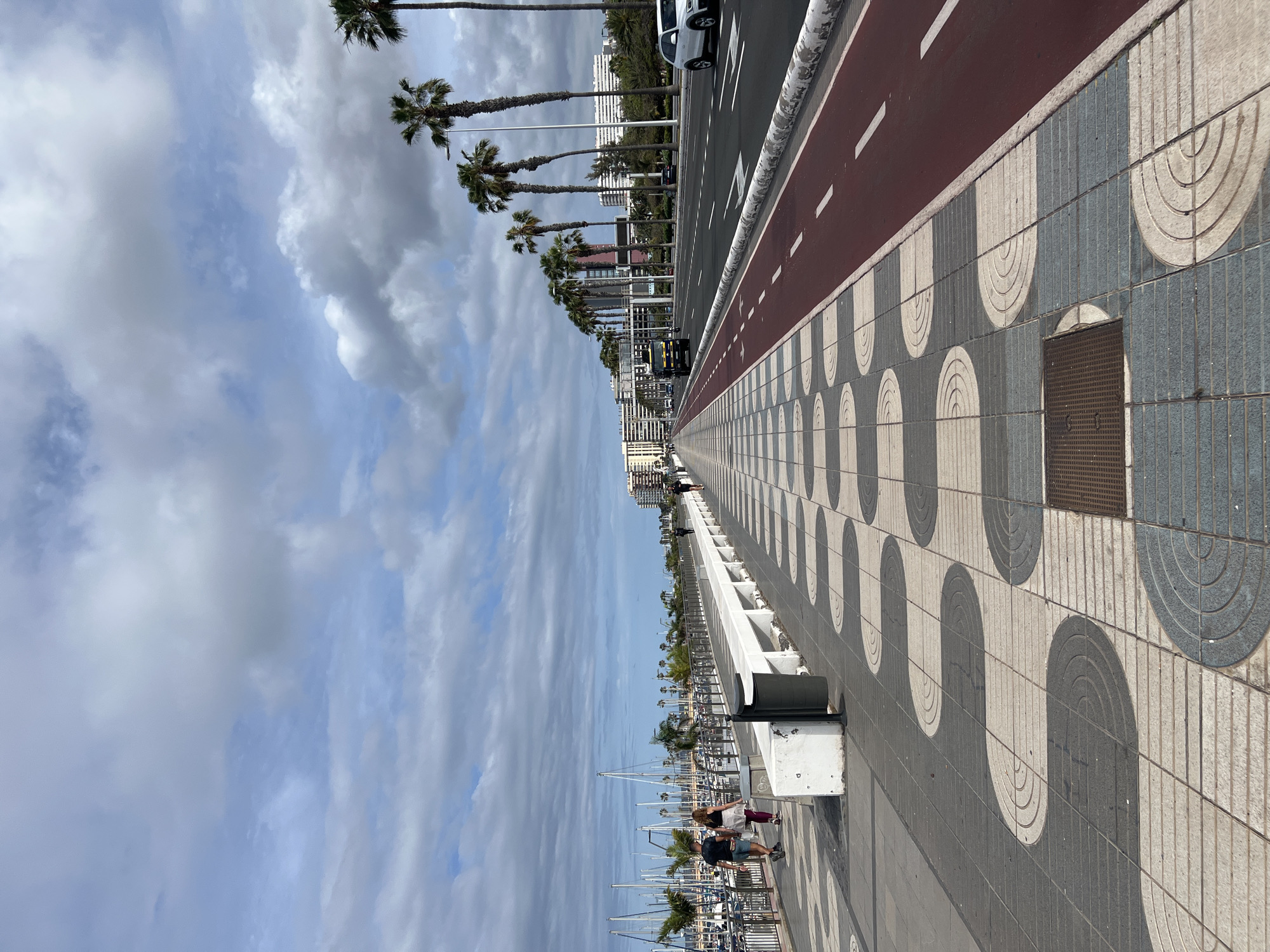 The image shows a view of the promenade at Las Palmas De Gran Canaria, Spain. The perspective is along a pathway paved with gray and white patterned tiles, with a distinct design featuring semicircular shapes. Adjacent to the pathway is a red bike lane.
Along the upper edge of the image is a road with palm trees lining the side, and a white car is parked.
On the left side, there's a view of a marina filled with boats. Two people are walking near a barrier along the marina. The sky is partly cloudy with patches of blue.