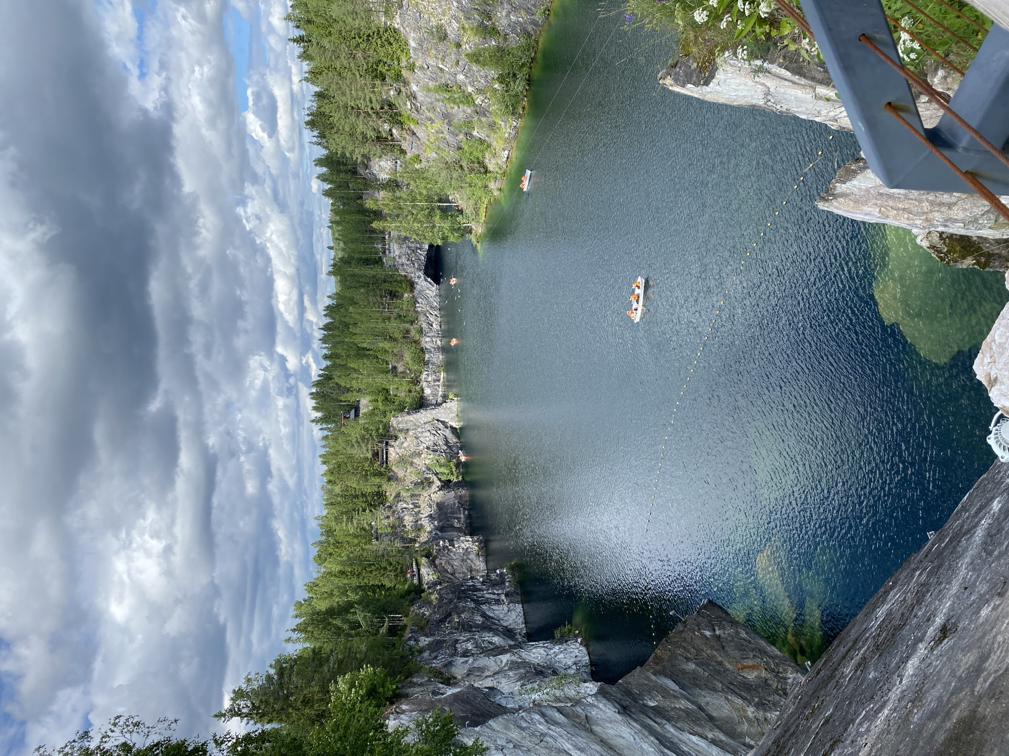 This is a vertical, high-angle photograph of Ruskeala Mountain Park in Russia. The scene depicts a quarry filled with clear, dark blue water, surrounded by steep, rocky cliffs and dense green forests. The sun is not directly visible, but the bright sky with dramatic, grey and white clouds suggests it is daytime, likely late morning or afternoon, and the weather is mixed with sun and clouds.
Several small boats, likely kayaks or canoes, are visible on the water, with people wearing orange life vests. One boat is in the middle distance, and another is closer to the shore on the left. There are also some small yellow buoys forming a line in the water.
The rocky cliffs display variations in texture and color, with some areas appearing smooth and others rough and fragmented. Green vegetation, including pine trees, grows abundantly on the cliffs and along the top edges. In the upper right corner, a metal railing and some white wildflowers are visible, indicating the vantage point is from a bridge or elevated walkway. The overall mood is serene and picturesque, highlighting the natural beauty of the location. No visible text is present in the image.
