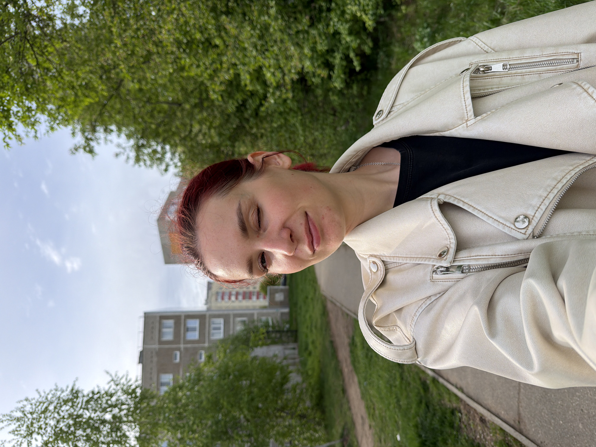 A reddish-haired person, with closed eyes and a slight smile, is seen from the chest up, wearing a white leather jacket over a black top. The photo is taken outdoors in what appears to be a park or green space. In the background is a concrete path, green grass, and trees with green foliage. Behind the trees, a light-colored apartment building is visible. Based on the architectural style and the environment, the photo appears to have been taken in a residential area of a city, possibly Chaikovsky, Russia as indicated by the prompt.