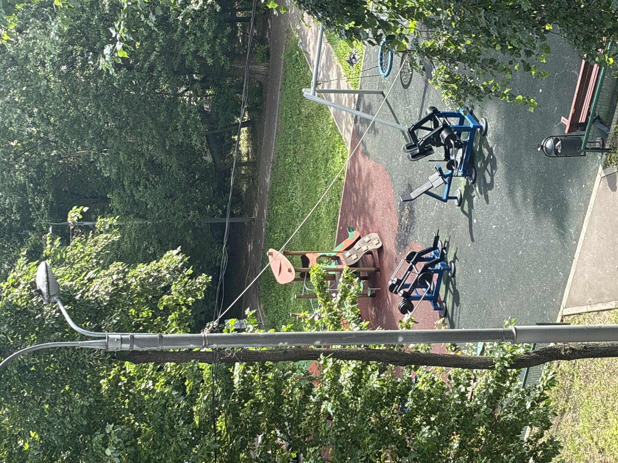 This is an aerial view of an outdoor fitness area and playground within a residential courtyard in Khimki, Russia, on a sunny day. A tall grey lamppost is prominently positioned in the foreground, partially obscuring the scene. The ground surface is a mottled green and red rubberized material, typical of a public recreational space.
The fitness area features two blue and black weight machines designed for leg presses or similar exercises. One machine is closer to the viewer, and the other is positioned further back and to the left. A trash receptacle and a bench with a red wooden seat and green metal frame are located near the edge of the fitness area.
Adjacent to the fitness equipment is a playground with a multi-colored climbing structure featuring slides and platforms. A swing set with a round blue net swing is also visible. The playground and fitness area are surrounded by dense green trees, indicating a lush, leafy environment. A paved pathway runs through the grass area behind the playground, and further back, more trees and a building are visible through the foliage. The overall impression is one of a well-maintained public space for recreation and exercise within an urban setting. No people are present in the image.