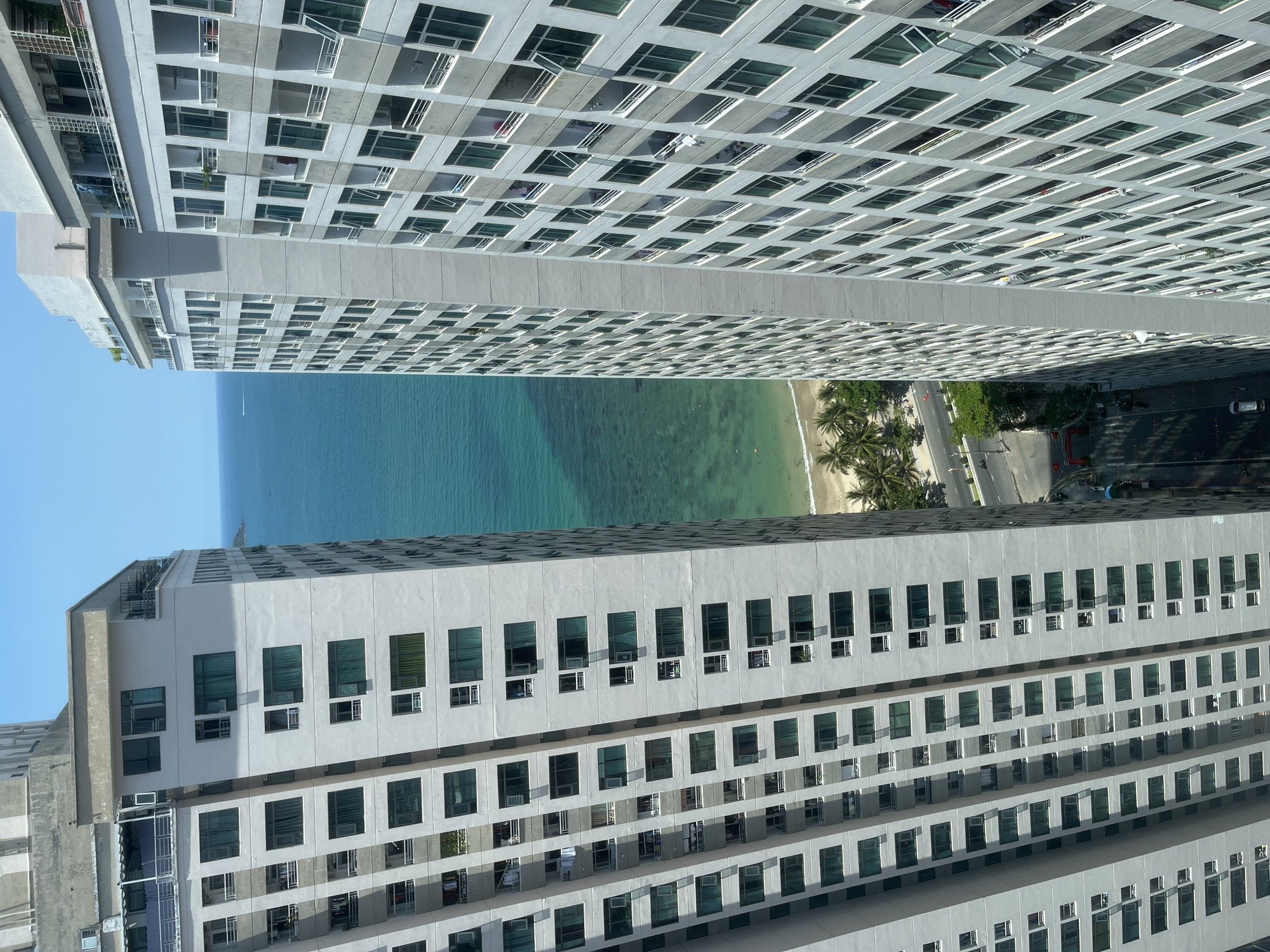 The image shows a view between two tall buildings in the city of Bac Nha Trang Ward, Vietnam. Visible between the buildings is a view of the beach, with light colored sand and clear water. A line of palm trees separate the beach from a paved road that runs along the shoreline. Cars and motorbikes can be seen on the road. The buildings themselves appear to be apartment complexes or hotels, with numerous windows and balconies.