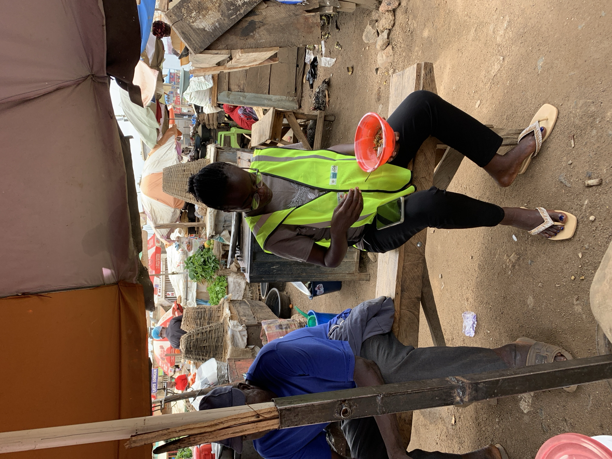 This media file depicts a lively outdoor market scene in Rafin Par, Nigeria, characterized by makeshift stalls and a dirt ground. The image is oriented sideways, but when viewed upright, it shows individuals engaging in daily activities under natural daylight.
In the mid-ground, a person sits on a simple wooden bench, wearing a bright yellow-green reflective vest, black trousers, and light-colored sandals. They hold a red bowl filled with food, possibly grains or rice, and a spoon, actively eating. A smartphone is placed beside them on the bench. The vest features a circular logo with blue and green elements, and the text "SOT" and partially "SUSTA..." (possibly "Sustainable") is visible.
Closer to the foreground, another individual is partially visible, also seated on a wooden structure. They wear a blue shirt, dark trousers, and dark sandals, appearing to be resting. A metal beam runs across the lower part of the frame.
The background reveals a bustling market environment with numerous stalls constructed from wooden planks and covered by various tarpaulins and fabric canopies in shades of brown, blue, white, and orange, offering shade from the sun. Among the market goods, woven baskets, sacks, and green leafy vegetables are discernible. On the dirt ground near the person in the vest, a small deceased bird, likely a chicken, is visible.
The scene is illuminated by natural daylight, indicating daytime. Faint signs in the distance include text like "Coca-Cola," "LISTS LONGER," and "PH," suggesting commercial advertising within the market. The overall atmosphere conveys the everyday rhythm of an informal marketplace.