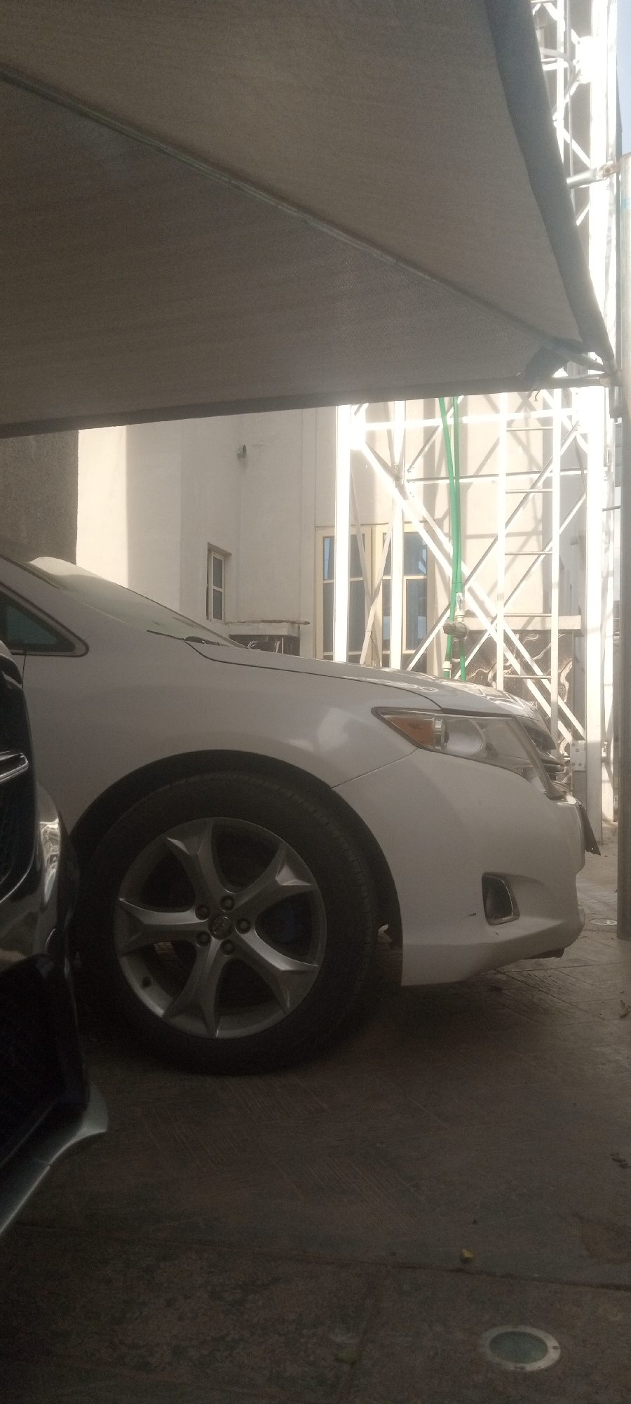 A white Toyota Venza is parked indoors under a shade structure in Narayi, Nigeria. The front passenger-side of the vehicle is visible, including the wheel, headlight, and front bumper. Part of another vehicle is visible on the left side of the image. Behind the Venza, a white building with windows is partially visible, along with a metal structure. The setting is indoors, under shade.