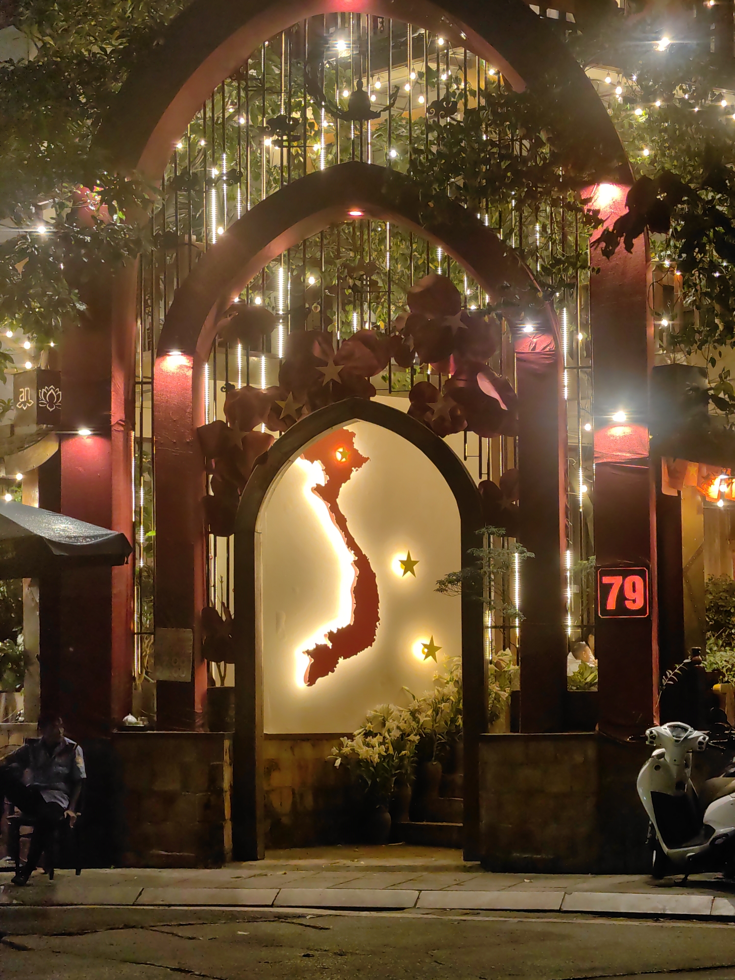 At night, the entrance of a building in Hà Nội, Vietnam is illuminated. An arched gate, decorated with a lighted outline of Vietnam, frames the entry. Strings of lights, along with greenery and red floral decorations, adorn the gate. To the left, a person sits in a chair. To the right, a scooter is parked in front of a sign that reads "79". The street is visible in the foreground.