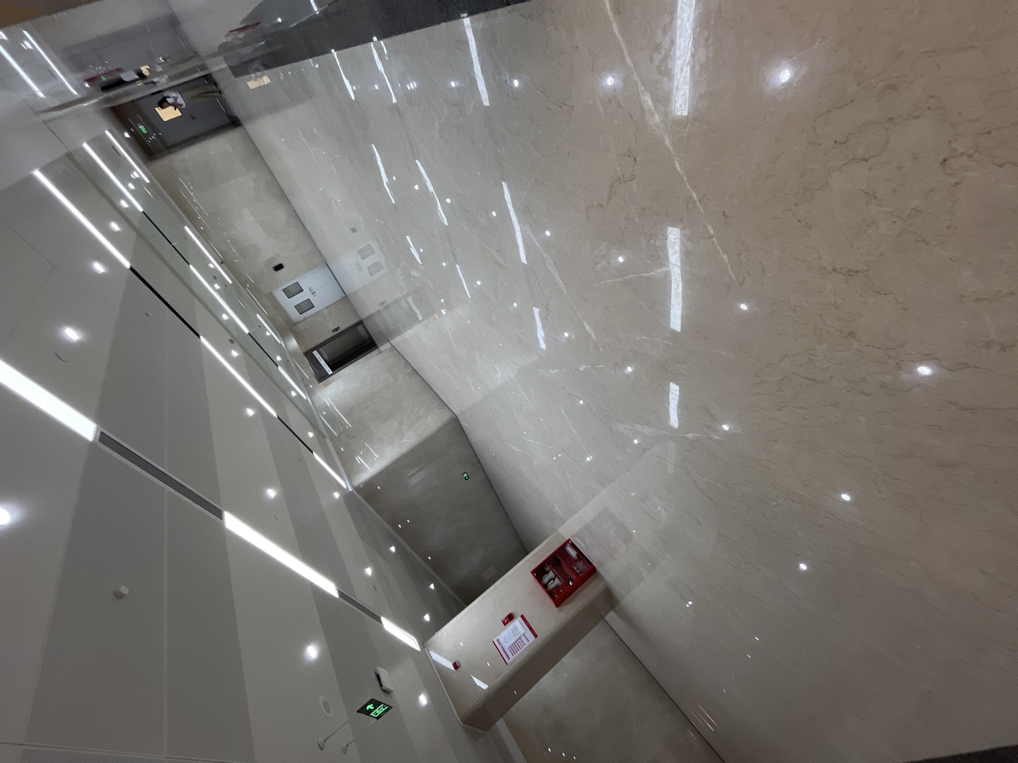 The image shows a well-lit, modern interior space, likely a commercial building or office hallway. The floor is made of polished beige marble tiles that reflect the overhead lights and the surrounding architecture. The ceiling features long, recessed fluorescent lights interspersed with spotlights. On the left side of the image, there is a pillar with a red fire extinguisher box mounted on it and a red and white sign above it. Further down the hallway, there is an elevator door. On the right side of the image, a person is visible in the background, standing near a doorway and looking at their phone. The overall environment is clean, spacious, and suggests a contemporary design. The location is Yuzhong District, China.