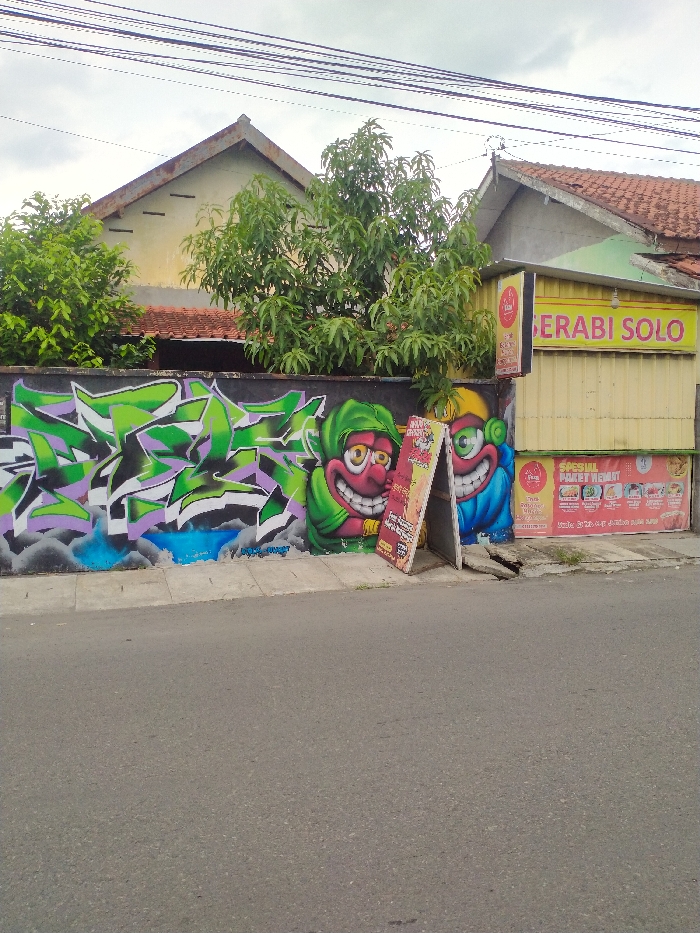 The image captures a daytime scene in Karanganyar, Indonesia, featuring a vibrant street-side wall and a food stall. In the foreground, a gray asphalt road stretches across the bottom, bordered by a concrete curb.
The main subject is a black wall adorned with colorful graffiti. On the left, large, jagged, abstract lettering in shades of green, purple, white, and black with blue highlights occupies a significant portion. To its right, two cartoonish characters are depicted. Both have large, wide smiles with prominent teeth and exaggerated, wide-open eyes. One character, predominantly green and red, wears a hood, while the other, mostly blue and yellow, also sports a hood.
In front of the graffiti, two A-frame sandwich board signs stand on the sidewalk, advertising various items. The text on these signs is largely unreadable, but one appears to promote a "DISKON" (discount) and "PROMO."
To the far right, a small, yellow-sided food stall or kiosk is visible. A prominent sign on its upper section reads "GERABI SOLO" in blue text on a yellow background. Below this, a banner advertises "SPESIAL PAKET HEMAT" (Special Economical Package) with images of food items and mentions "THIS FRIDAY & SATURDAY."
In the background, behind the wall and stall, are parts of residential houses with reddish-brown tiled roofs. Lush green trees are interspersed between the buildings and the wall. Overhead, multiple black power lines crisscross against a cloudy, overcast sky, indicating a bright but not sunny day. No people are visible, and there is no specific activity taking place in the captured moment.