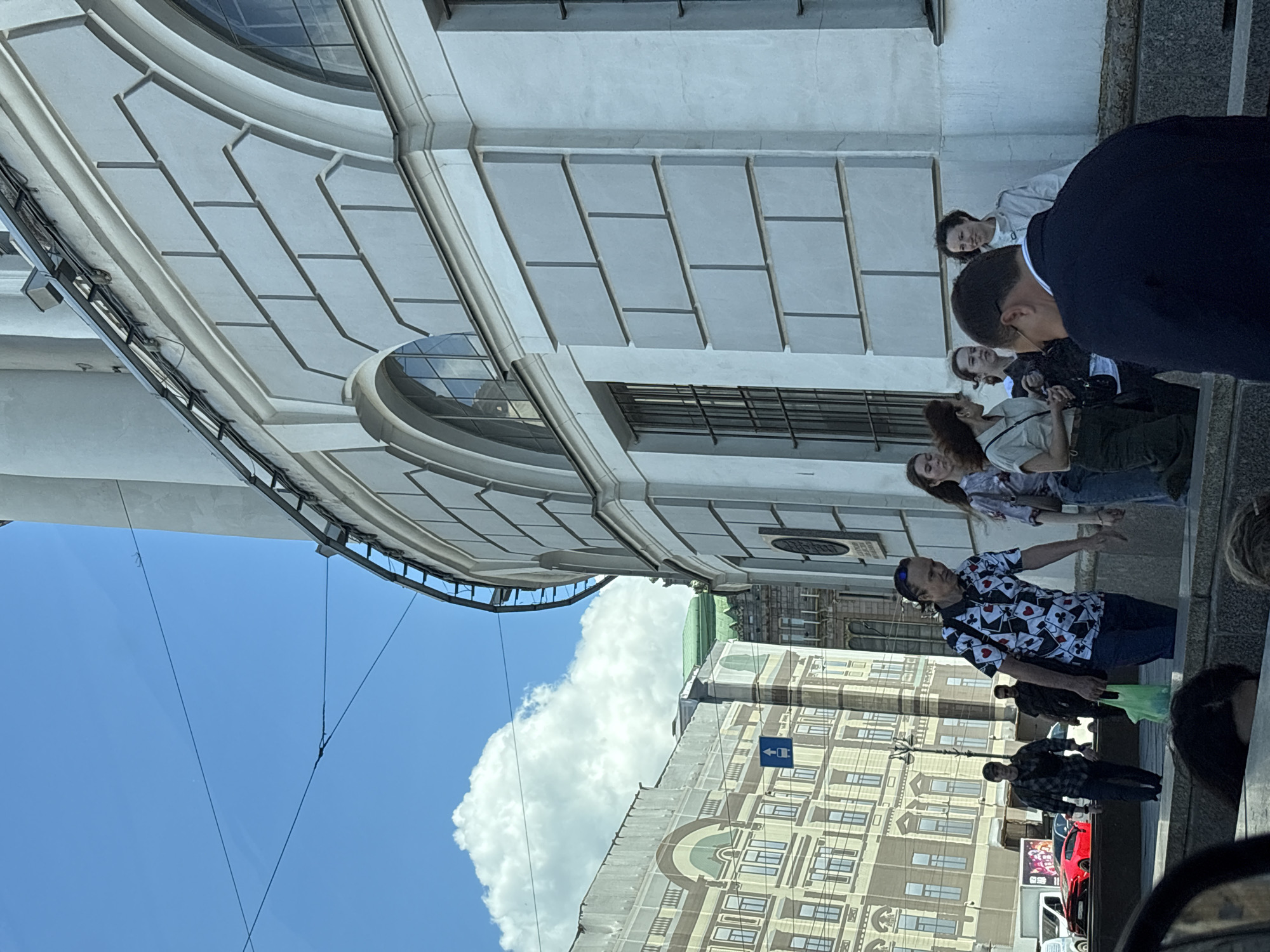 The image captures a street scene in Saint Petersburg, Russia, with a prominent neoclassical building on the right side of the frame. The building features a light gray facade with decorative stone accents and arched windows. A group of people are visible walking on a sidewalk in front of the building, some interacting with each other. Among them, a man in a black suit is seen from the back, facing away from the camera. To the left, another building with a detailed facade and arched windows is partially visible, suggesting a historic district. The sky is blue with fluffy white clouds, and some overhead wires are present. A red sports car is parked further down the street. The overall atmosphere appears to be a typical daytime urban environment.