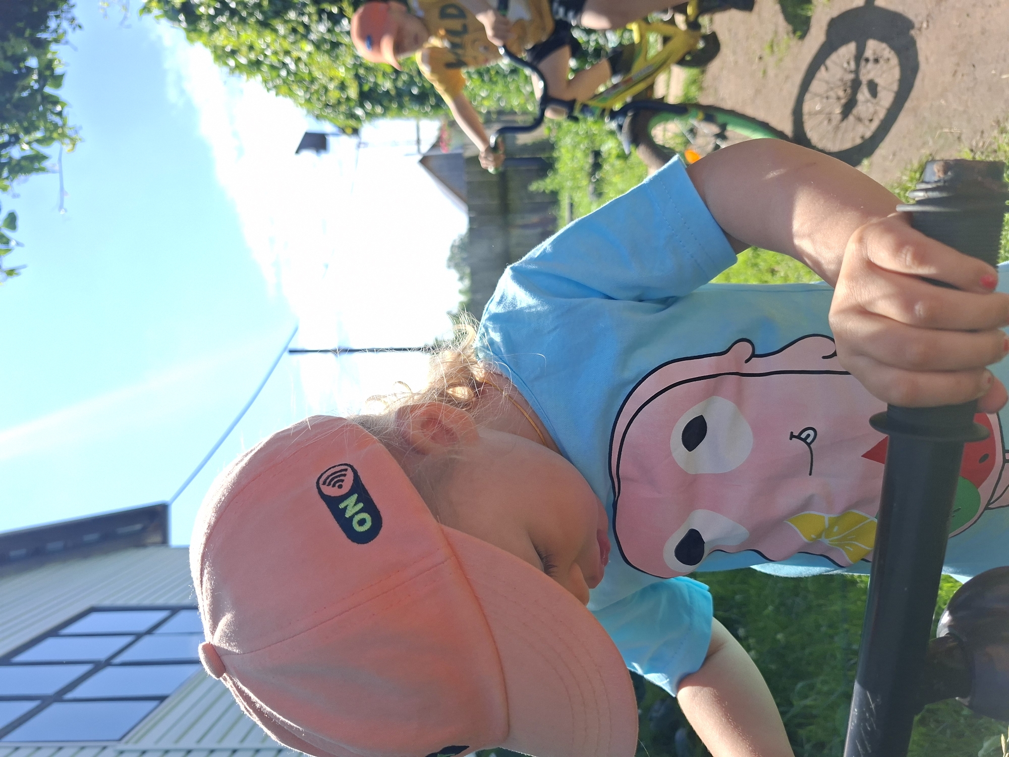 This is a medium shot of two young children outdoors on a sunny day, riding bicycles. The foreground is dominated by the left side of a child wearing a light blue t-shirt with a cartoon print and an orange baseball cap with an "ON" logo. The child is gripping the handlebars of a bicycle. In the background, another child wearing a yellow t-shirt and an orange cap is riding a yellow bicycle. The environment appears to be a backyard or park area with green grass, dirt paths, and some trees and foliage. The scene is set in the city of Karsha, Russia.
