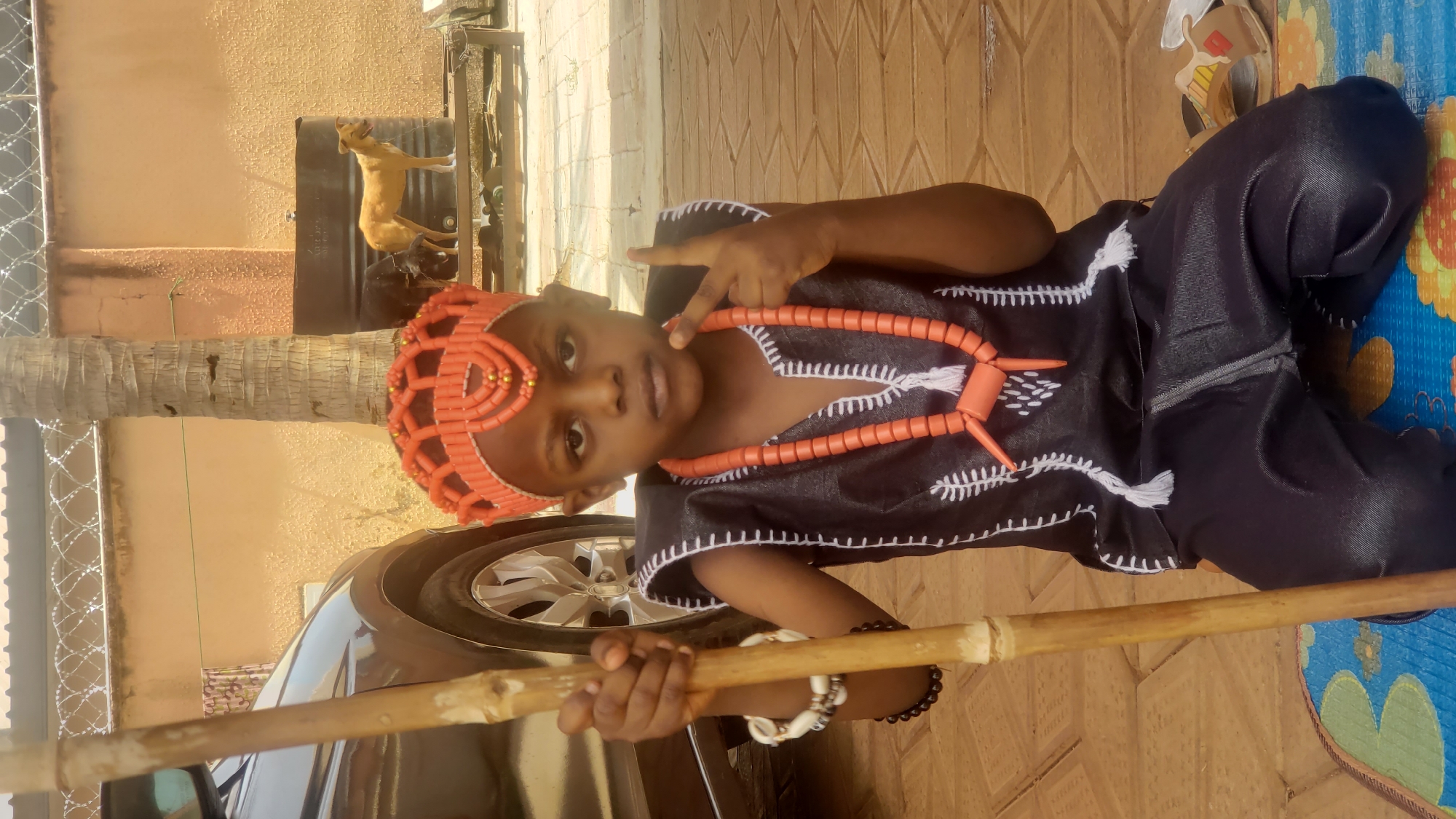 A child in traditional attire poses outside in Sabon Tasha, Nigeria. The child wears a black outfit with white fringe and an orange beaded necklace and headwear. They hold a wooden stick and make a peace sign. A black car, a palm tree, a metal barrel with a dog on top, and a building are visible in the background. The ground is paved. A mat lies on the right side of the frame.