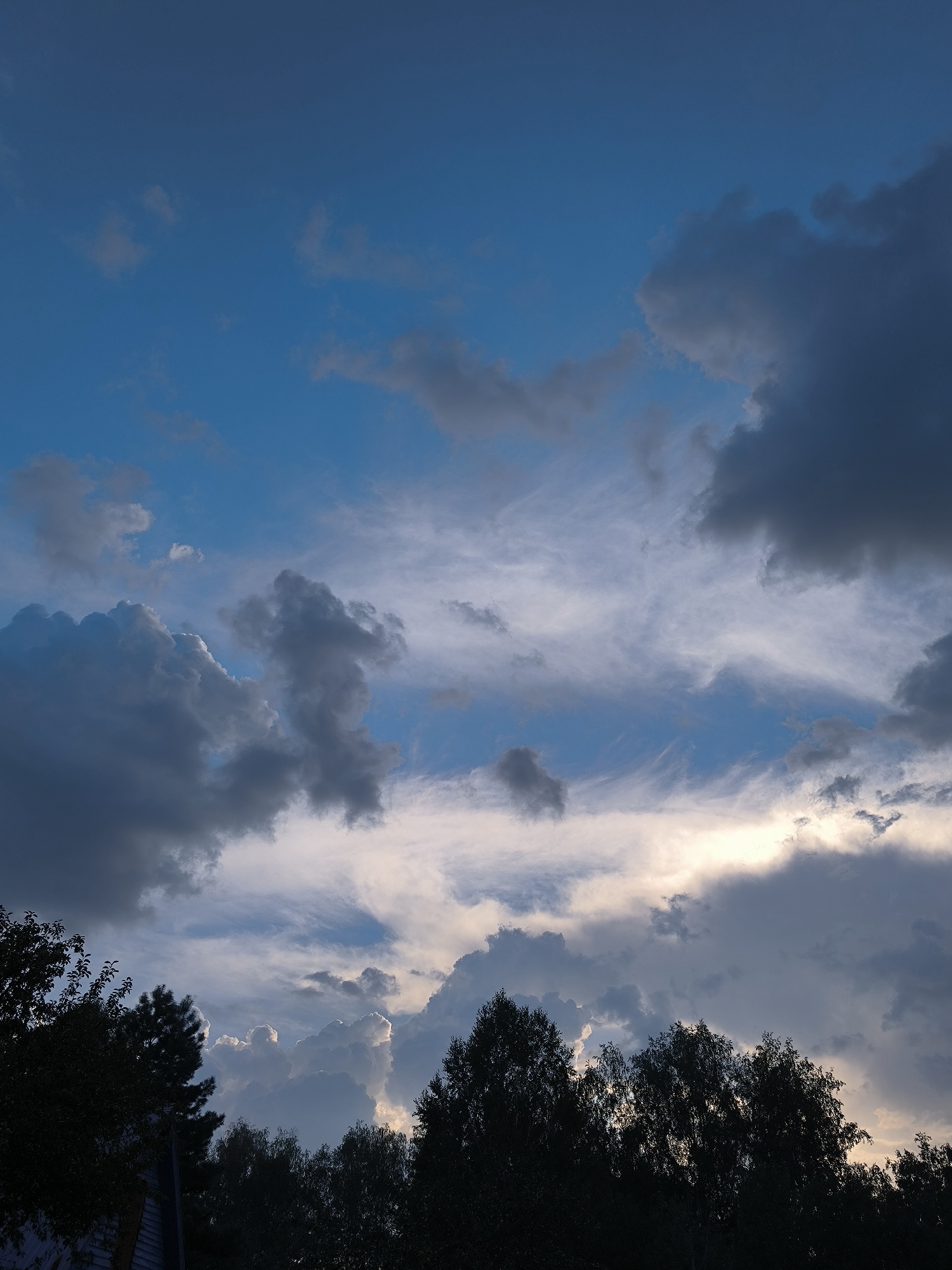 A low-angle shot captures a dramatic sky filled with a mix of dark, heavy clouds and wispy, white cirrus clouds against a deep blue backdrop. Sunlight filters through a gap in the clouds, creating a bright, ethereal glow. The silhouette of a line of trees runs across the bottom of the frame, partially obscuring a building that appears to be a house. The overall impression is one of an approaching storm or the aftermath of one, with the interplay of light and shadow creating a sense of depth and movement. The location is the city of Maiskii, Russia.