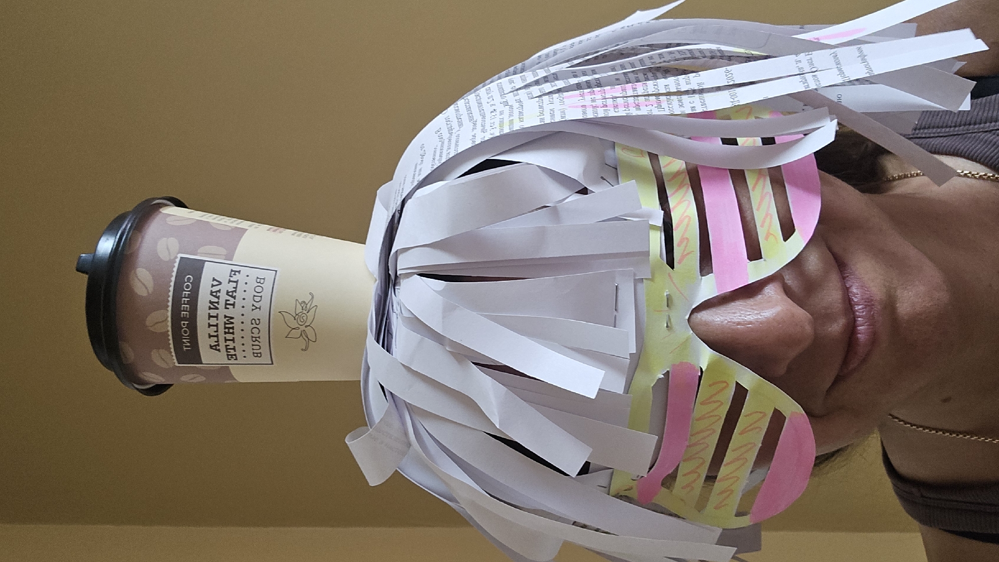 A person wears a homemade costume. The costume includes a coffee cup worn as a hat, made to look like it says "Coffee Point" and "Vanilla Flat White Body Scrub". The hat is adorned with paper strips that resemble hair. In front of the eyes, the person wears oversized, colorful glasses made of paper, with yellow and pink stripes. The background is a plain, muted yellow wall. The image appears to be taken indoors.