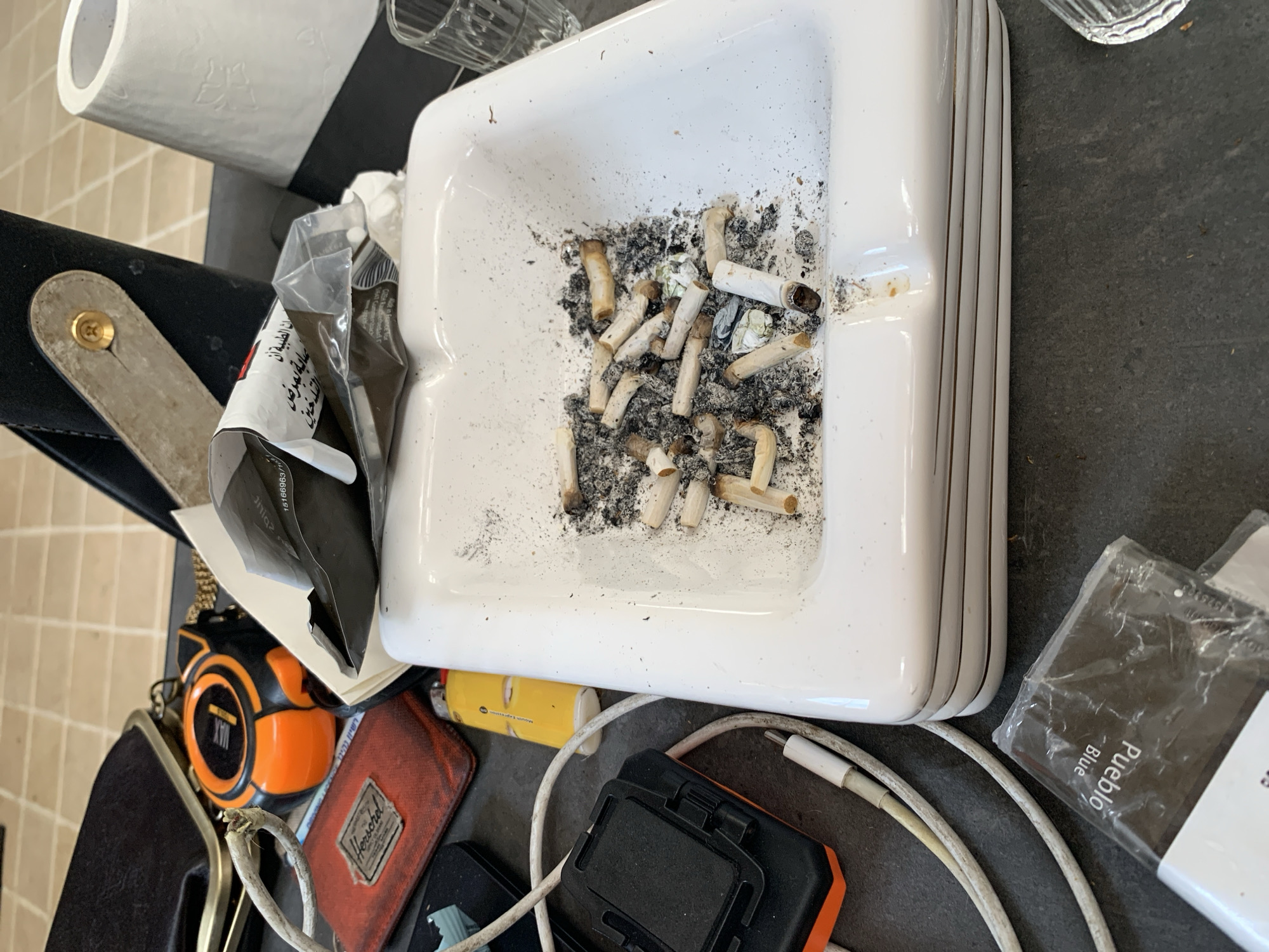 The image captures an overhead view of a cluttered surface, likely a table or countertop, filled with various personal items, dominated by a large white ceramic ashtray. The ashtray is heavily used, containing numerous cigarette butts and a substantial amount of ash.
To the left of the ashtray, a roll of white toilet paper sits on a black holder. Below it, a black and white package with Arabic text is visible, part of which reads "عملية تمريض". Further down, an orange and black tape measure, with "MAX" visible on its side, is placed next to a red wallet displaying the "Herschel" brand logo. A black purse or wallet with a metallic clasp is also present.
Below the ashtray and extending to the right, a yellow disposable lighter is partially visible, with "Mouth expression" possibly printed on its side. Several white charging cables are coiled and intertwined around a black rectangular electronic device. In the bottom right corner, a clear plastic-wrapped package of rolling tobacco is visible, labeled "Pueblo Blue". A clear glass is also partly seen in the top right corner of the frame. The background includes a tiled surface, possibly a floor or wall, in a light, neutral color. The overall scene suggests a casual, perhaps unkempt, indoor environment. The presence of the ashtray, cigarette butts, and rolling tobacco indicates recent or ongoing smoking activity in this area.