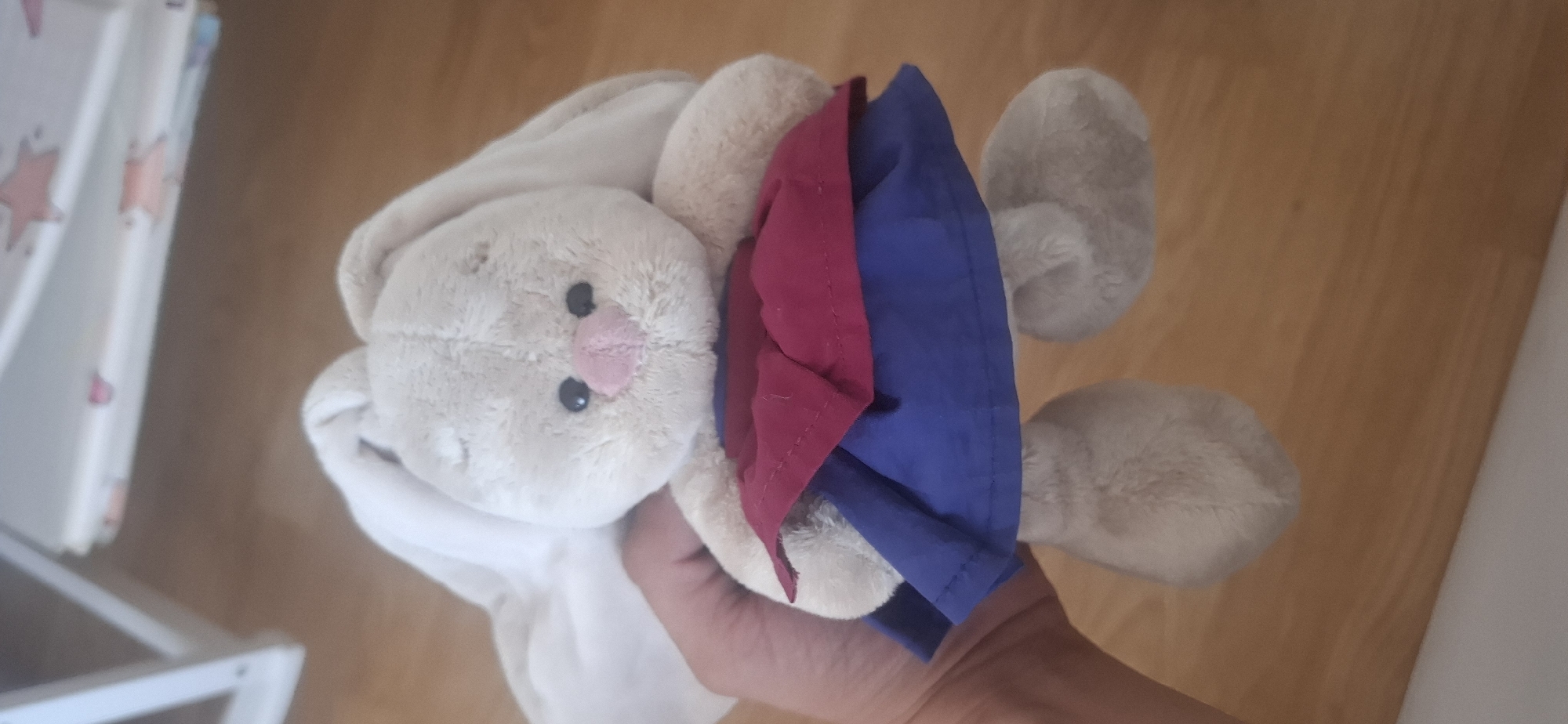 A light beige plush bunny toy with long ears is held in a person's hand. The bunny is wearing a two-layered skirt, with a red layer over a blue layer. The bunny has black button eyes and a pink nose. The background consists of a wooden floor and a blurry white surface with some pink star shapes. The image was taken in Ust'-Katav, Russia.