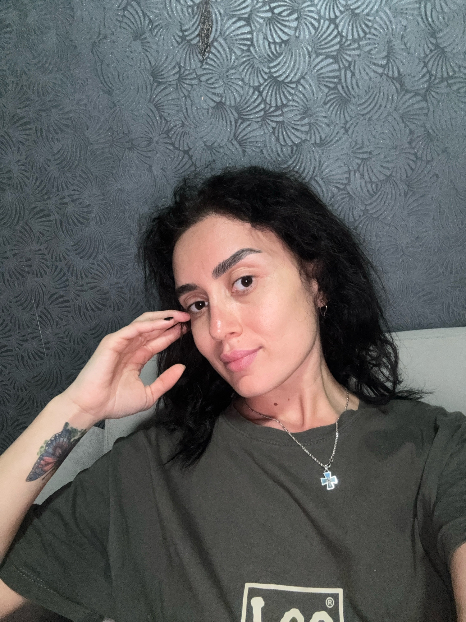 A woman with dark, curly hair is shown from the chest up, looking towards the camera. She is wearing a dark green t-shirt and a silver necklace with a cross pendant. A butterfly tattoo is visible on her left arm. She is touching her face with her right hand, near her eye. The background is a dark grey textured wallpaper with a repeating fan-like pattern. The scene appears to be indoors, possibly in a bedroom, with a white headboard visible on the right side of the image. The lighting is soft and comes from the front, casting a slight glow on her face. There is no indication of the location being in Tbilisi, Georgia.