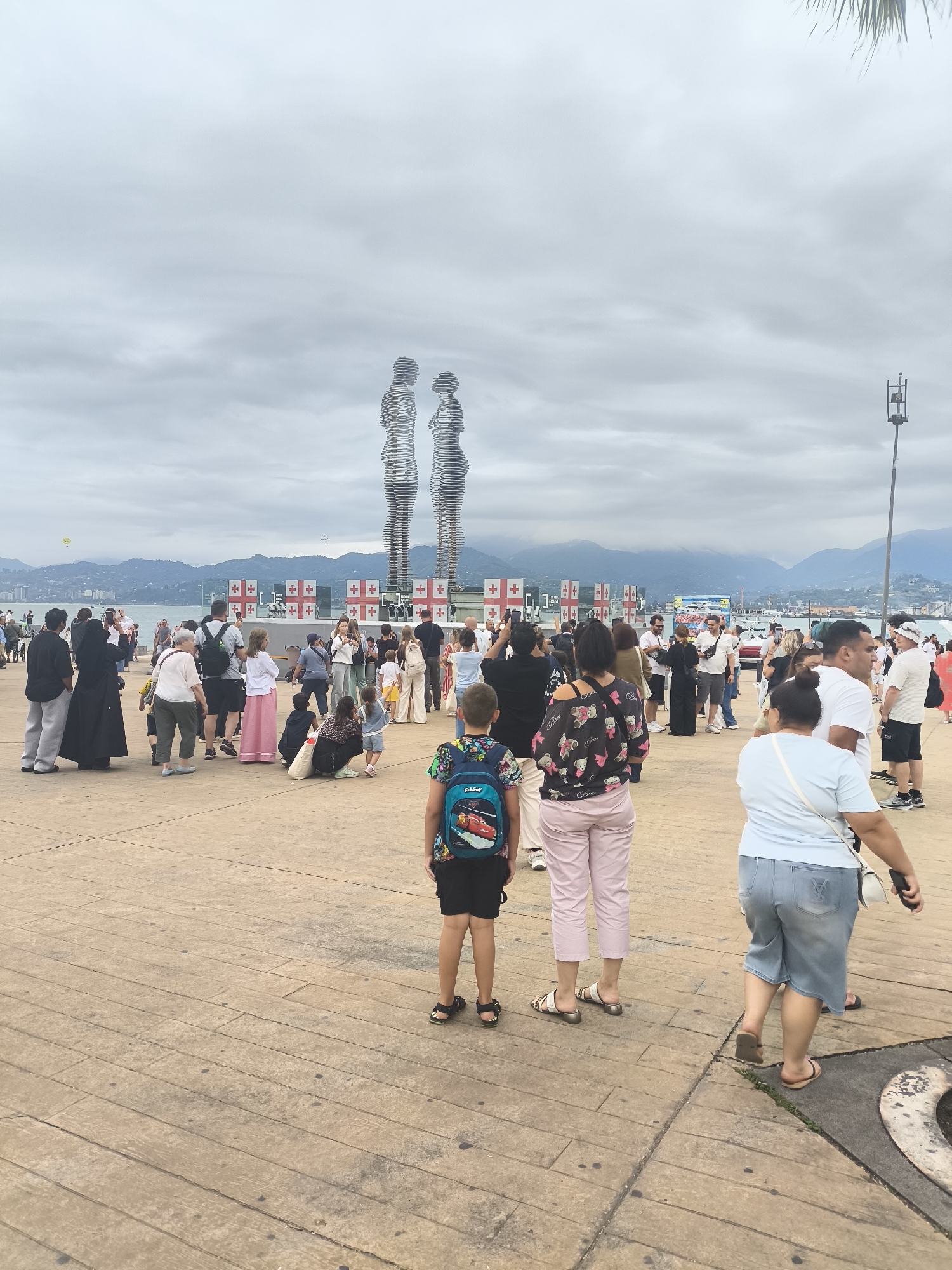 In Batumi, Georgia, a crowd of people gathers on a paved promenade near the Black Sea. Two large, abstract metallic sculptures of human figures, known as the Ali and Nino statues, stand prominently in the background, facing each other. Behind them, a series of flags or banners with a red cross on a white background are displayed. The sky is overcast, and a range of mountains is visible in the distance. People of various ages are present, some walking, others standing and observing. A young boy wearing a backpack with a cartoon character is in the foreground. The promenade is made of concrete slabs.