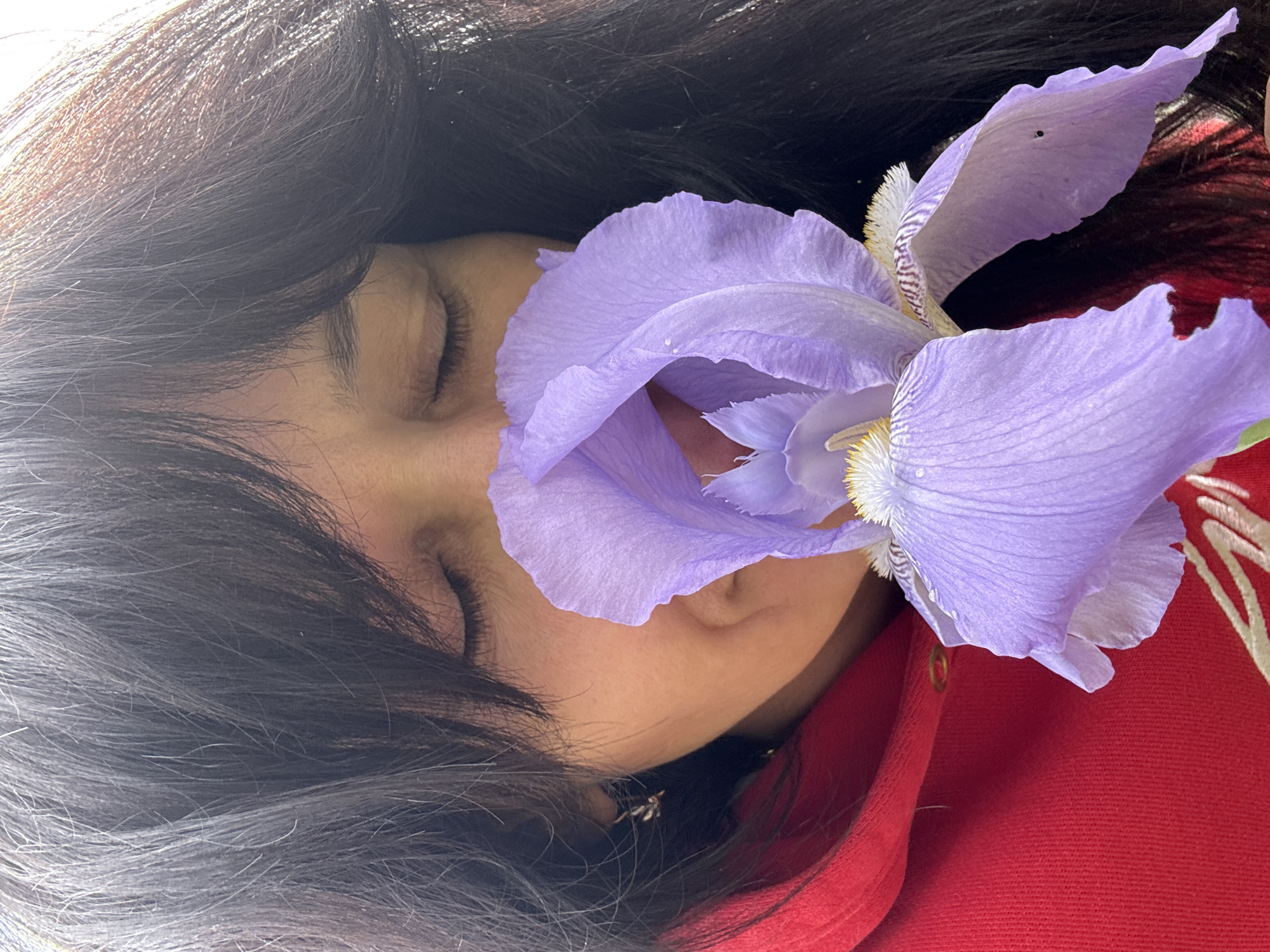 A close-up shot features a person with dark hair and closed eyes. A large purple iris flower is positioned near their face. They are wearing a red hoodie. The exact location cannot be determined from the image.