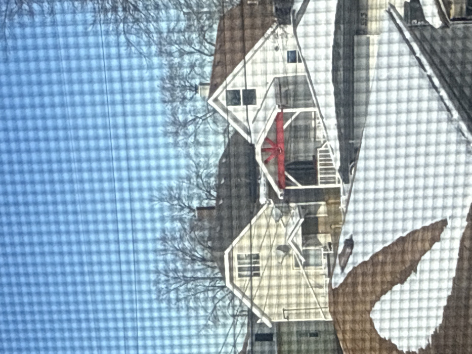 Two houses with gabled roofs stand side by side under a clear, blue sky. The houses feature white trim, including railings and stairs, with one displaying a red, wheel-like structure. Bare tree branches are visible in the background. A portion of a larger building, possibly a barn or larger house, is visible on the right, its roof covered with a white material, possibly snow.