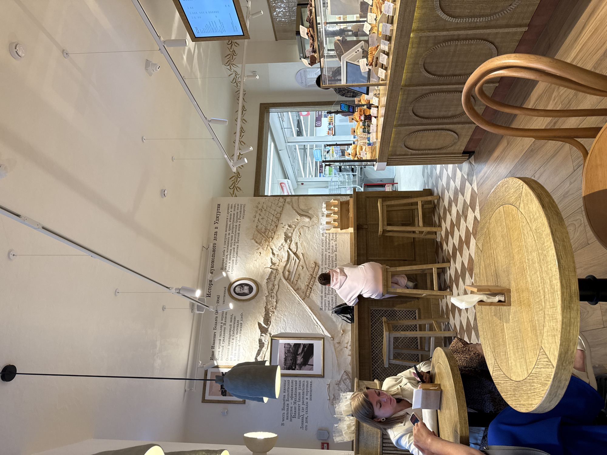 This is an interior view of a modern bakery and cafe in Izhevsk, Russia. The scene is dominated by warm, natural wood tones, with checkered flooring in the foreground and wooden plank flooring in the background.
In the foreground, two round wooden tables are visible, each with a single chair. The table on the left has a woman sitting at it, holding her phone and looking down. The table on the right has a napkin dispenser.
The midground features a counter with various baked goods displayed under glass. Behind the counter, there is a wall decorated with historical murals and text, depicting local history. Two individuals are seated at a bar area in front of this wall, one facing away from the camera, wearing a pink hoodie, and another partially visible from the side.
The background shows an entrance to a supermarket, with bright lighting and shelves visible. Overhead, a track lighting system illuminates the space, along with pendant lights. The overall ambiance is that of a cozy yet contemporary establishment.