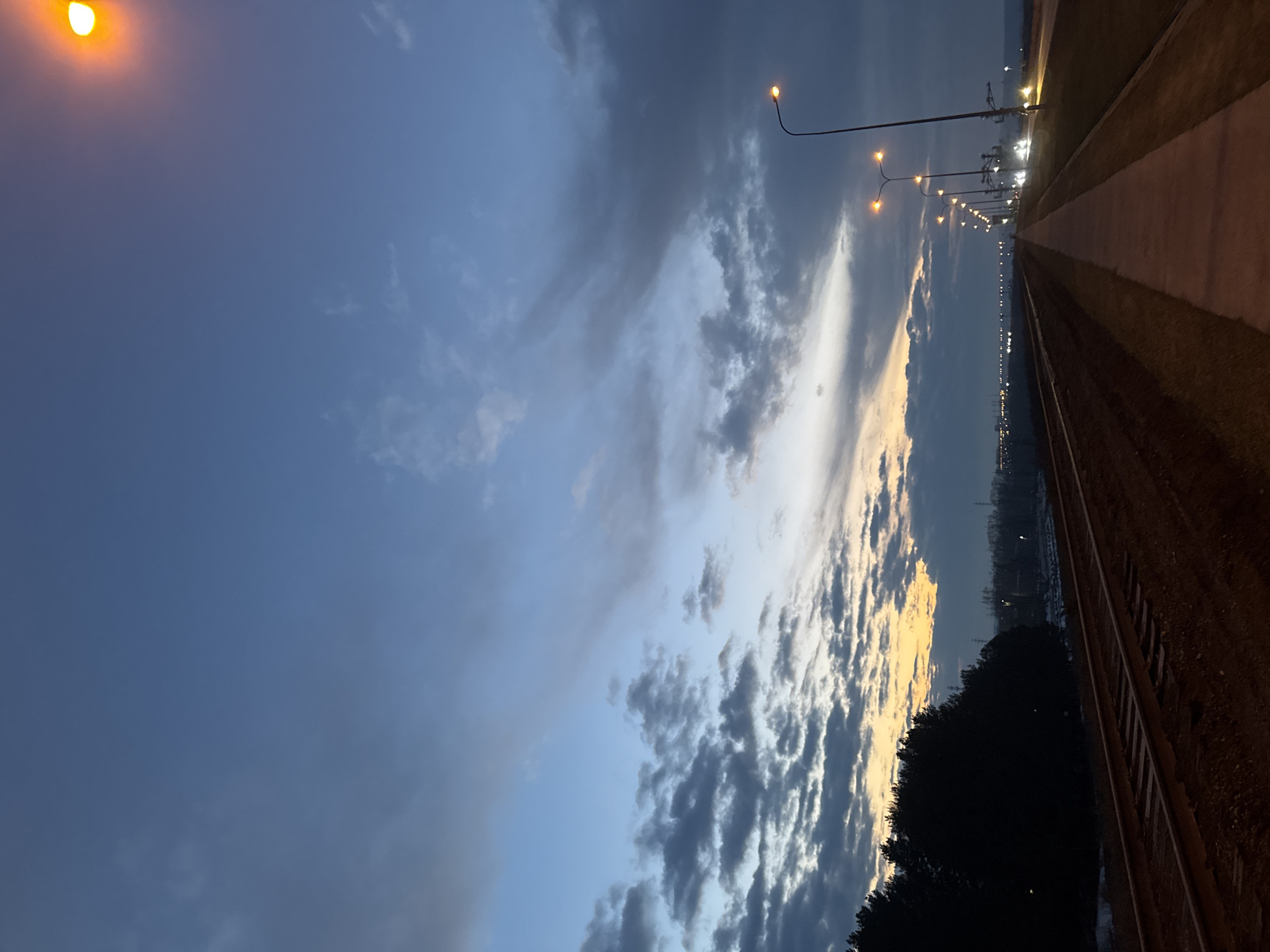 The image depicts an evening scene in Chaikovsky, Russia. The sky transitions from a deep blue on the left to a twilight hue on the right, with dark, textured clouds illuminated from below by a fading sunset. A row of street lamps casts a warm glow along a path or road, which is lined with what appears to be a track or railway. Dark vegetation is visible in the foreground. The overall impression is one of dusk settling over an urban or suburban landscape.