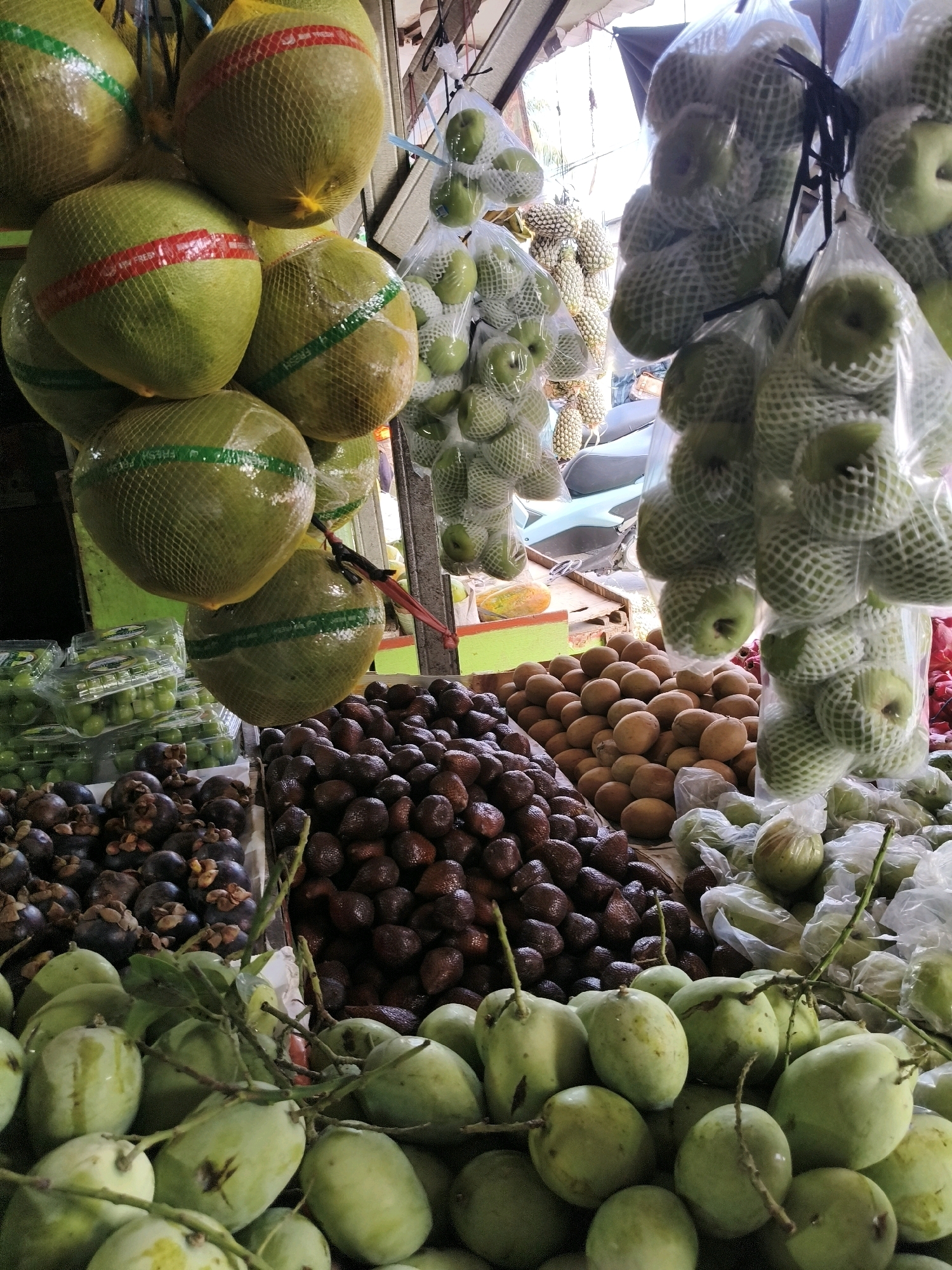 A street market stall in Bandung City, Indonesia, displays an abundant variety of fresh fruits. In the foreground, a large pile of green mangoes, some with stems still attached, are partially obscured by a tangle of thin branches. Behind the mangoes, to the left, are dark purple mangosteens, and to the right, rows of salak fruit, also known as snake fruit, with their distinctive scaly brown skin. In the middle ground, more salak fruit and piles of sapodilla are visible. Hanging from above and to the left, large green pomelos are individually wrapped in protective netting, some adorned with red and green bands. To the right, similar netting-wrapped fruits, likely guavas or small apples, hang in bunches. Further back, more packaged fruits and glimpses of motorcycles suggest a busy urban market environment. The stall's roofing and supporting structures are visible, indicating an open-air setup. The overall scene is a vibrant and colorful arrangement of produce, typical of an Indonesian fruit market.