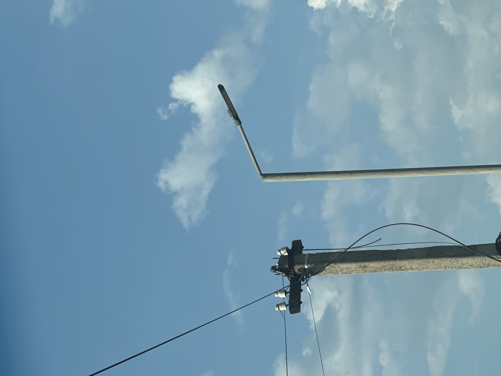 In Shatava, Ukraine, an overcast sky with scattered white clouds provides the backdrop for a low-angle shot of two utility poles. To the left, a concrete pole stands tall, supporting electrical wires and ceramic insulators. A darker, thinner wire extends diagonally from the upper left corner of the frame, crossing over the top of the pole. To the right, a lamppost with a curved arm and a dark fixture stands against the sky. The lamppost appears weathered, its surface textured with rust or discoloration. The scene suggests an outdoor setting, possibly on a street or in a rural area, under a daytime sky.