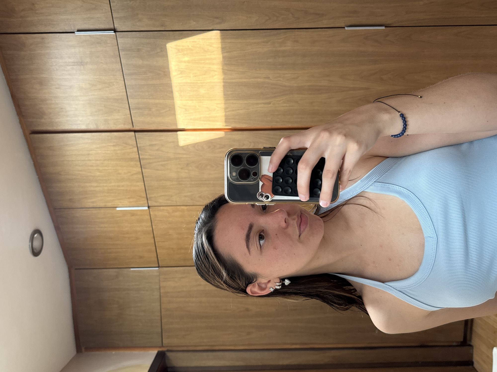 A person is taking a selfie in front of a wooden cabinet. The person is wearing a light blue tank top and has a bracelet on their left wrist. The cell phone has a case with a cartoon character and black circular grips on the back. The location appears to be indoors, with a wall and a light fixture visible on the left side. There are no details to indicate the city of San Bernardino Tlaxcalancingo, Mexico.