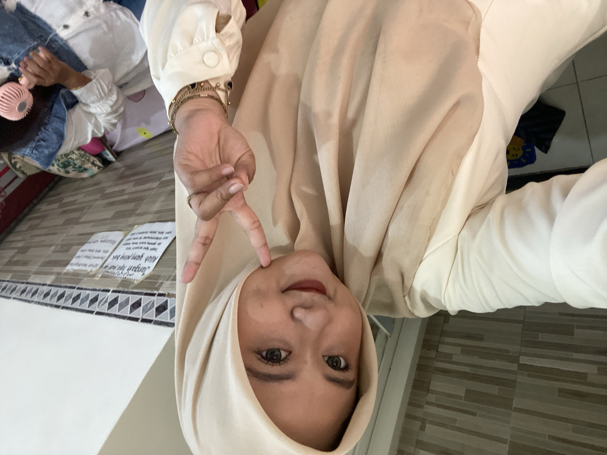 A woman with brown eyes is indoors in Jember, Indonesia. She is wearing a light beige hijab and top. Her right hand is raised, gesturing a peace sign near her face. Her left hand and arm are visible in the upper left corner. There are two stickers on the wall behind her near the left edge of the frame. The floor is tiled. A small child, wearing blue and white clothes, can be seen behind her in the upper left corner playing with a pink fan.