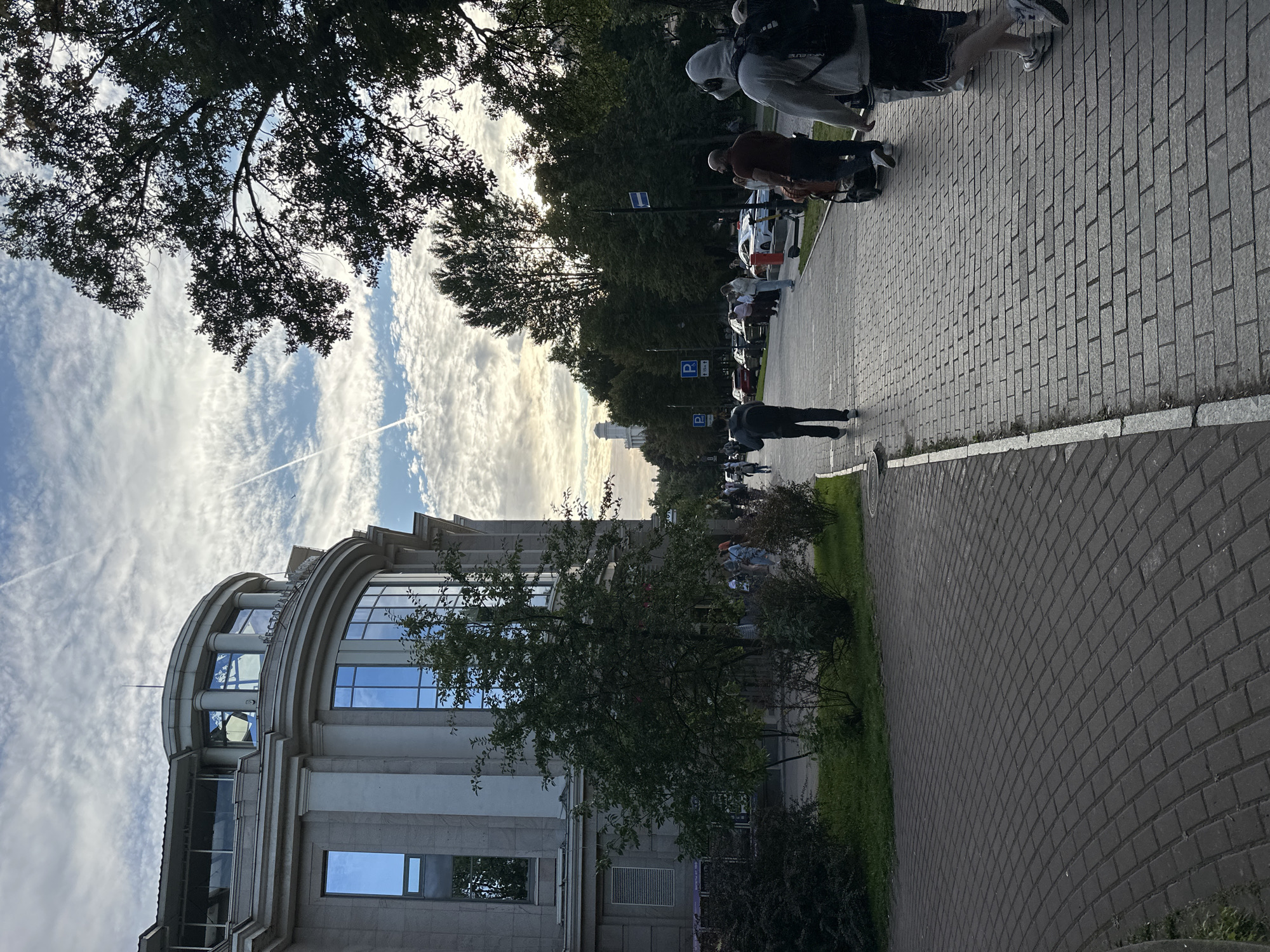 A paved pedestrian walkway lines a street in Saint Petersburg, Russia, with a large, modern building on the left and a line of parked cars and trees on the right. The sky above is cloudy with visible contrails. Several people are walking along the path, some with backpacks. One person is pushing a stroller, and another is on a scooter. The building appears to be a commercial establishment, possibly with offices or shops, featuring large windows and a curved architectural element. The street is lined with trees, and there are parking signs visible. The overall atmosphere is that of a public urban space during daylight hours.