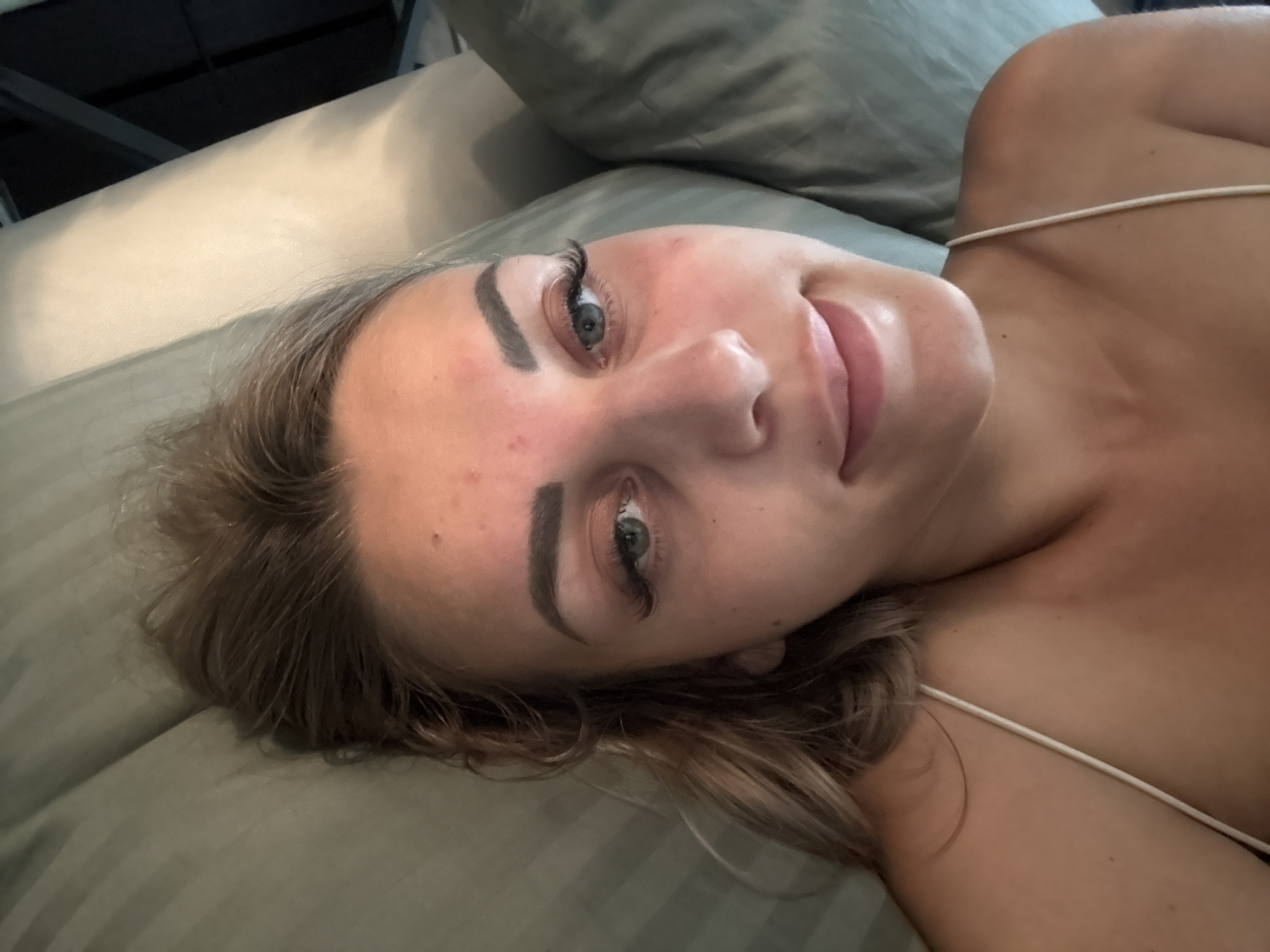 A young woman with light brown hair and green eyes lies on a green cushioned surface, possibly a sofa or bed. She is wearing a thin strap, likely from a tank top or bikini. Her face is tilted, and she is looking upwards and slightly to her left. Her makeup includes mascara and what appear to be false eyelashes, and her lips are painted with a light pink lipstick. The background consists of soft green cushioning and a darker object above. The scene appears to be indoors.