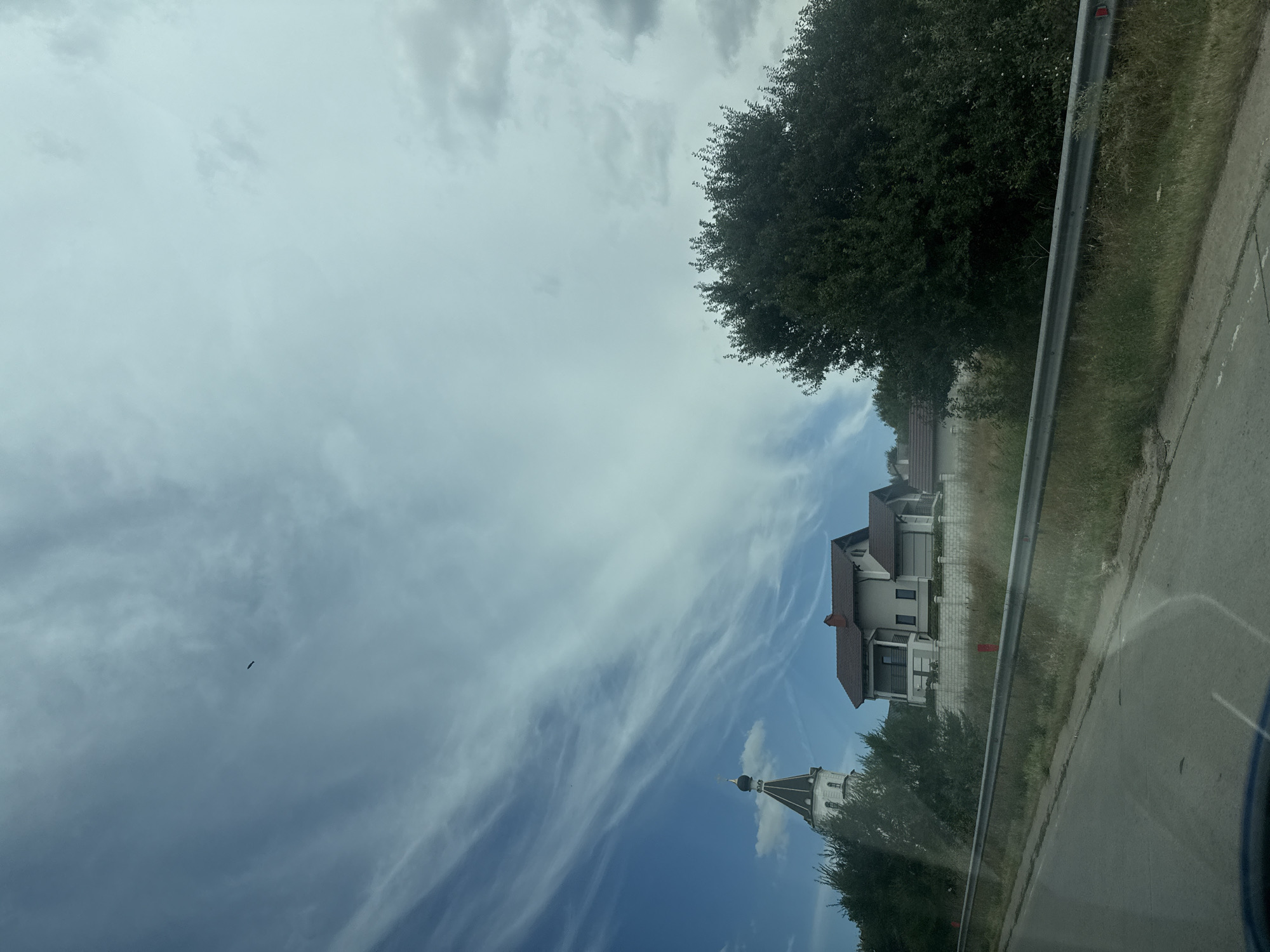 The image shows a view from a moving vehicle on a road in Băcioi, Moldova. On the left side of the frame, a white church tower with a dark dome and a cross on top is visible, partially obscured by trees. Adjacent to the church, a modern two-story house with a light-colored facade and a red-tiled roof is situated on a slight incline. The house has multiple windows and balconies. Further to the right, a large, dense green tree dominates the right side of the image. A metal guardrail runs along the side of the road, separating it from the grassy embankment where the houses and trees are located. The sky is partly cloudy with streaks of cirrus clouds and patches of blue sky. The road surface is asphalt, and there is dry grass and some vegetation on the embankment. The overall scene depicts a semi-rural or suburban area on a cloudy day.