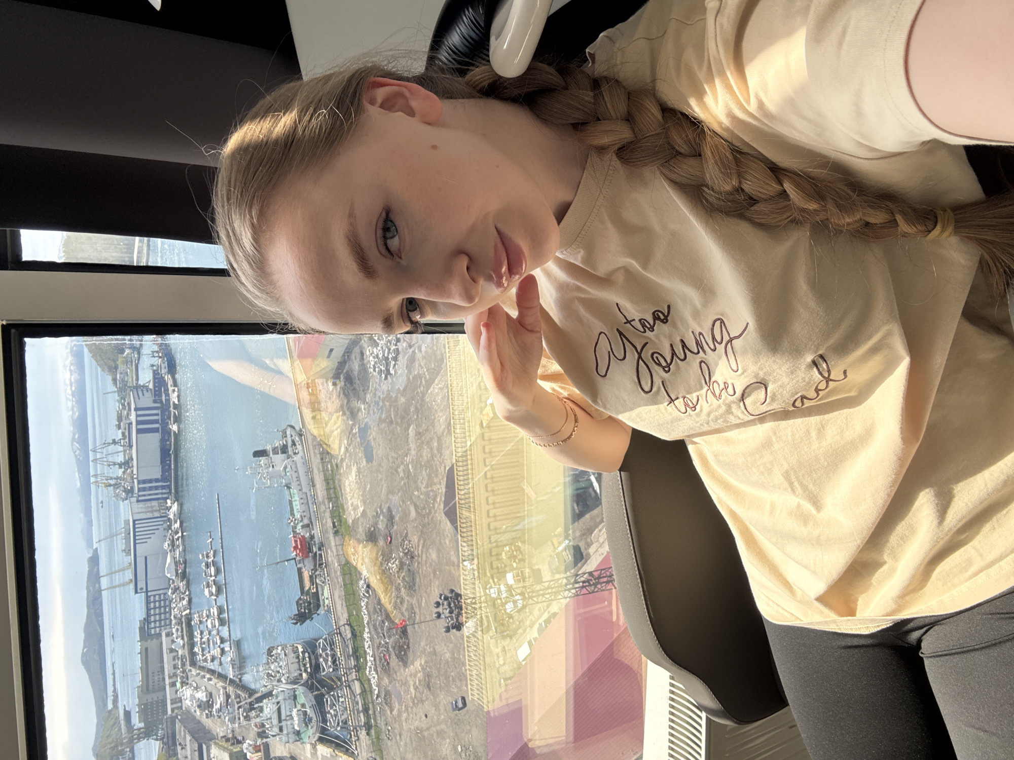 A young woman with long, light brown hair styled in a thick braid is captured in a selfie. She is wearing a light beige t-shirt and dark leggings, seated in what appears to be a black office chair. Her expression is calm, and she looks directly at the camera, with one hand gently touching her chin. A thin, multi-strand gold bracelet is visible on her right wrist.
The image is taken from a high vantage point, looking out through a large window that offers a panoramic view of an industrial port city, identified as Petropavlovsk-Kamchatsky, Russia. The sunlight is bright, suggesting a clear day, possibly morning or afternoon, with strong highlights visible on her hair and the scenery outside.
The extensive view outside the window showcases a wide body of water, likely a bay, filled with numerous ships, including large cargo or industrial vessels docked at piers, and many smaller boats. The shoreline features various industrial buildings, some with blue and white facades, alongside cranes and what appear to be construction or storage areas. In the distance, mountains or hills rise under a clear sky. Reflections of interior elements or nearby buildings are subtly visible on the window glass, indicating the height of her location.
On her t-shirt, stylized script is visible, spelling out the phrase "too Young to be Sad."
