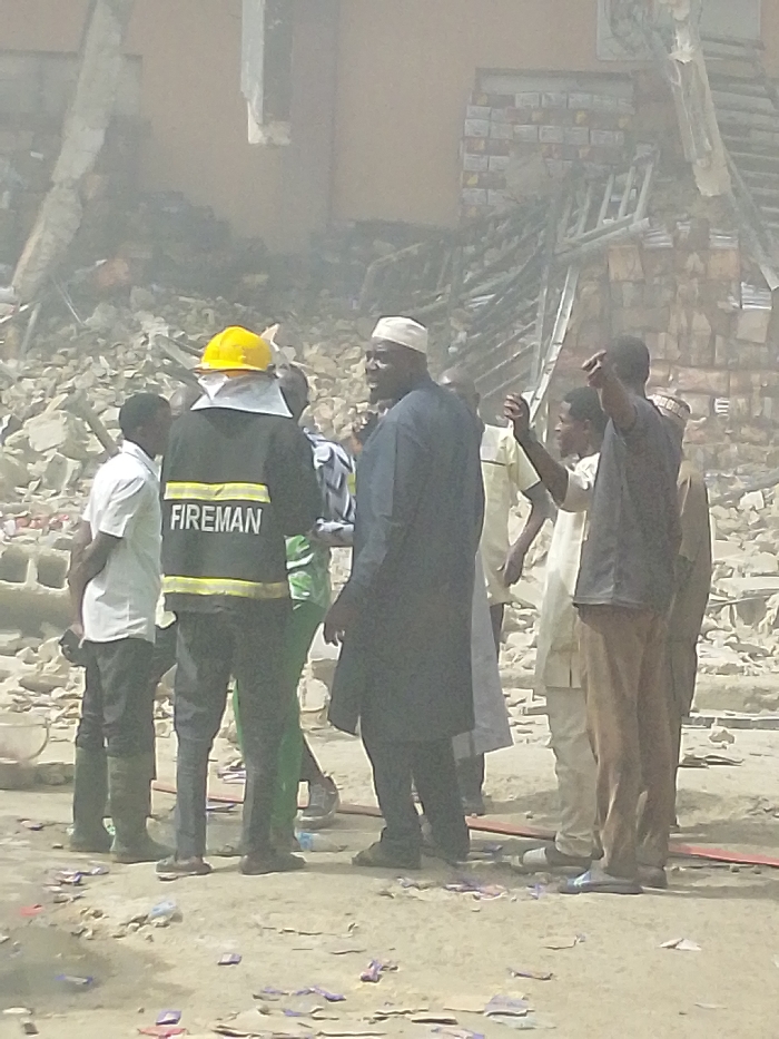 The image depicts a post-incident scene in Fagge C, Nigeria, characterized by extensive structural damage and debris. In the foreground, a group of approximately eight to ten men is gathered on uneven, dusty ground littered with small pieces of debris. A man wearing a black "FIREMAN" jacket with yellow lettering, a yellow hard hat, and a white neck protector stands with his back mostly to the camera, positioned slightly left of center. To his right, a man in a dark traditional tunic and white skullcap faces left, appearing to communicate or observe. Another man to the right of the individual in the dark tunic has his right arm raised.
The background is dominated by a large pile of rubble, consistent with a collapsed or severely damaged building. Visible elements within the debris include fractured concrete blocks, twisted metal, and sections of brick wall. Portions of a damaged wall structure and a bent ladder or scaffolding element are identifiable within the rubble on the right side. The entire scene is enveloped in a visible haze, suggesting dust or smoke, and is under daylight conditions.