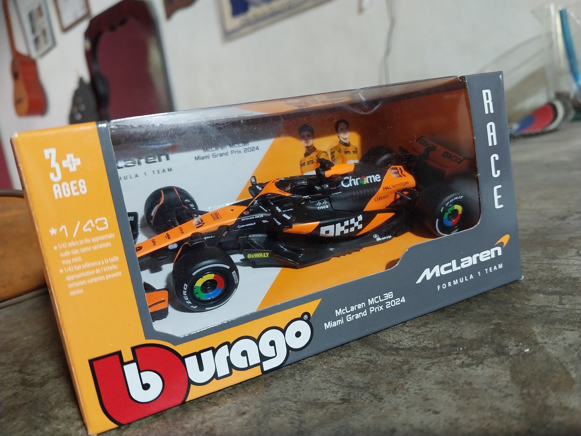 The image depicts a McLaren MCL38 Formula 1 Team diecast model car, still packaged in its original box, located in Tinaquillo, Venezuela. The car is orange and black, featuring sponsor logos. The box is primarily yellow and gray with the "Burago" logo prominently displayed. "RACE" is written vertically on the box's side. The box indicates the car is a 1/43 scale model of the McLaren MCL38 from the Miami Grand Prix 2024. The background is somewhat blurred, showing glimpses of furniture and objects within the room. The surface the box is sitting on appears to be a rough, possibly concrete, floor.