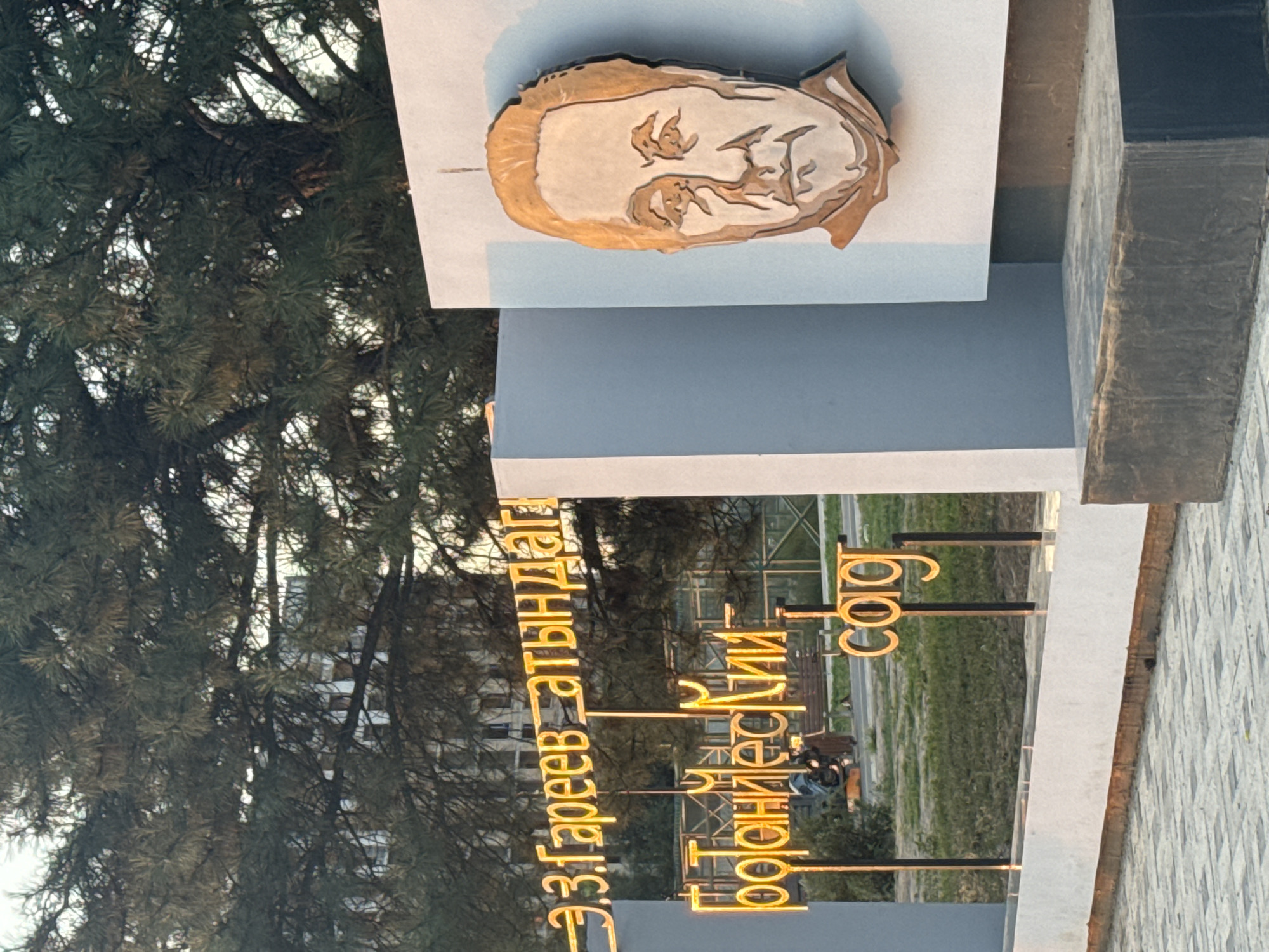The image depicts an outdoor scene featuring a monument with a portrait of a man and illuminated signage. The portrait appears to be a relief sculpture of a man's head and shoulders, rendered in a metallic material, mounted on a light-colored vertical surface. To the left of the portrait, there is illuminated signage in a golden-yellow neon font, displaying Cyrillic text. The text is arranged in multiple lines and appears to be a dedication or a name. In the background, a large tree with dense foliage is visible, partially obscuring a building with multiple windows. The ground in front of the monument is paved with interlocking square tiles. A low-lying grassy area is also visible behind the signage. The overall lighting suggests it is either early morning or late afternoon. The monument is located at the entrance to the Bishkek Botanical Garden, which is named after E.Z. Gareev.