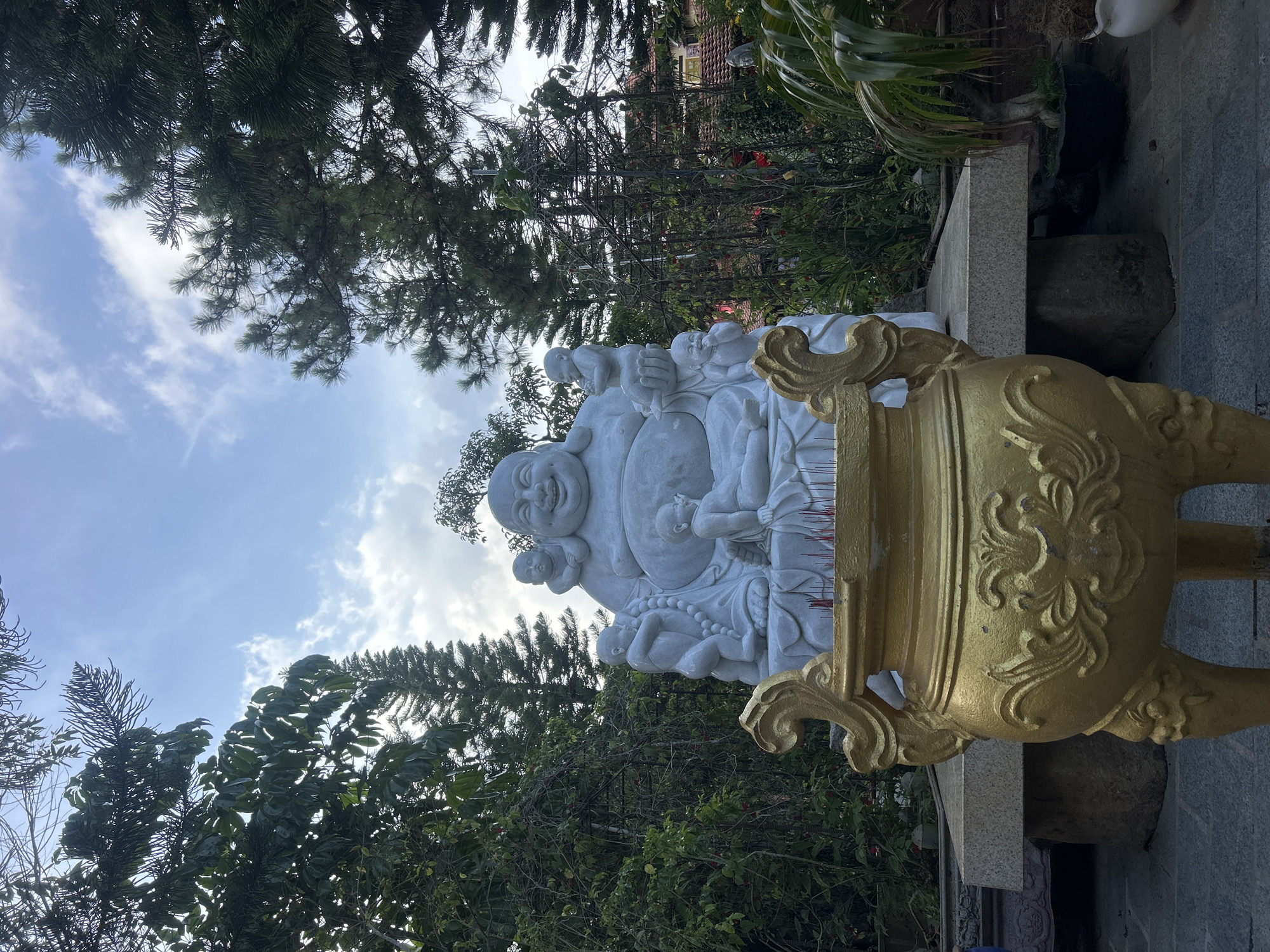 A mid-level, horizontal shot of a white Buddha statue beside a gold colored urn, located in the city of Lâm Viên - Đà Lạt, Vietnam. The statue is smiling and surrounded by smaller figures. It is placed on a stone base, and the urn is positioned to the right. The background shows trees and a blue sky with white clouds.