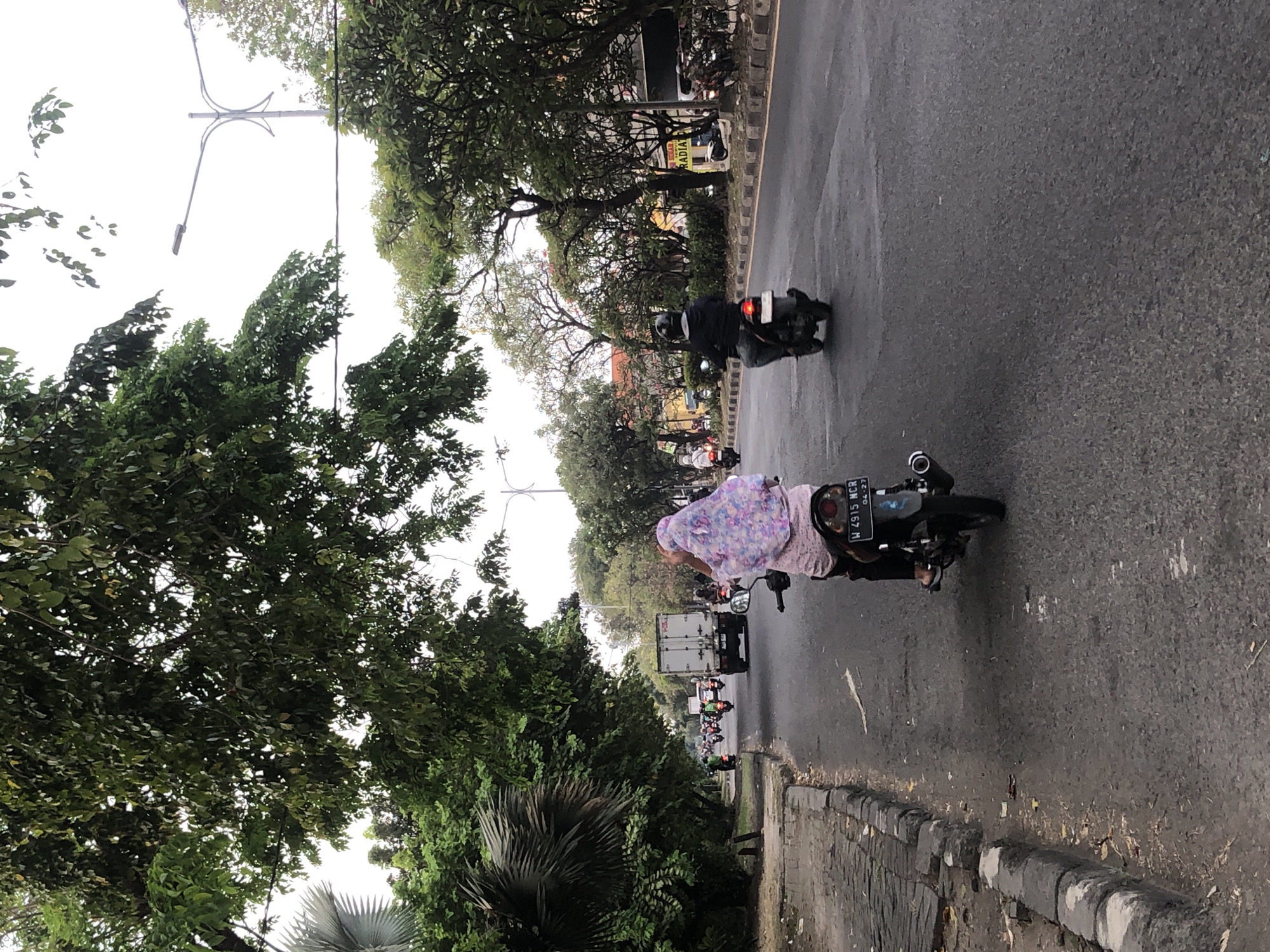 The image captures a street scene in Sumorame, Indonesia, with a focus on traffic and the urban environment. A woman wearing a colorful headscarf rides a motorcycle in the foreground, her back to the camera. She is followed by another motorcyclist, a man wearing a black jacket and helmet. In the distance, a white truck and numerous other motorcycles are visible, indicating a moderate amount of traffic. The street is lined with trees on both sides, providing shade and a natural element to the urban landscape. Streetlights and some buildings can be seen in the background, suggesting the presence of a developed area. The road surface appears to be asphalt, with a curb and sidewalk visible on the left side of the image. The sky is overcast, suggesting a diffused light source. The overall impression is that of a typical day in a bustling Indonesian city.