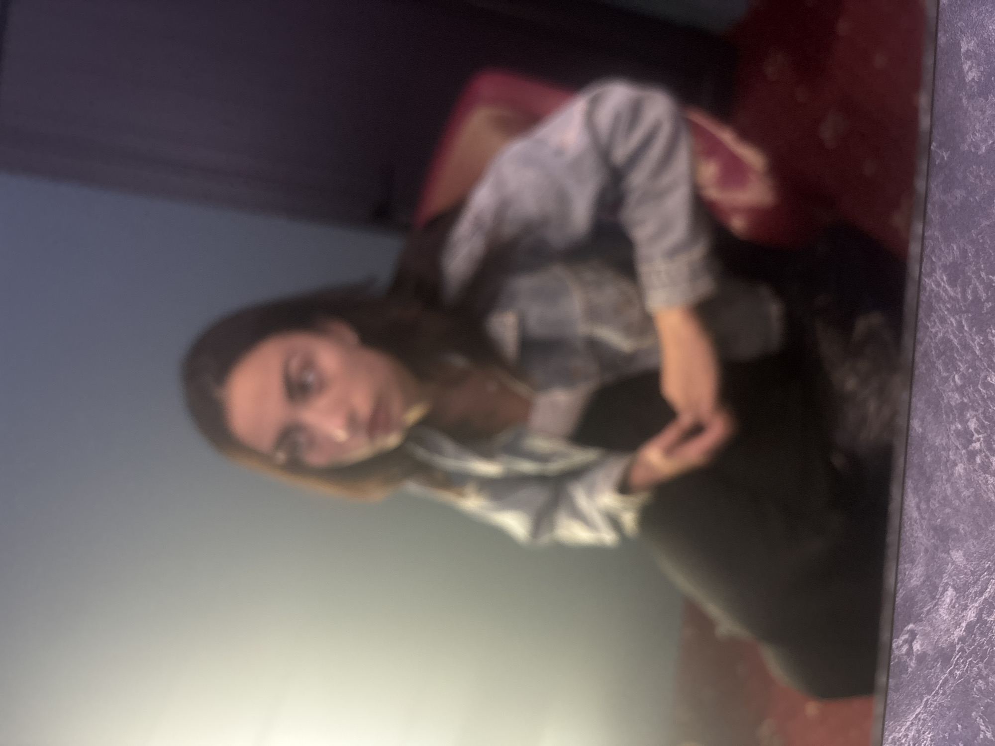 A young woman with long, dark hair is seated in a chair. She is wearing a light blue denim jacket over a white top and black pants. Her hands are clasped in her lap. She is looking off to the side, away from the camera. The chair she is sitting in appears to be upholstered in a deep pink fabric with a patterned texture. The background is dimly lit with a bright light source to the left, casting a soft glow. To the right, a dark doorway or wall is visible. The floor is covered with a patterned carpet in shades of red and brown. The image appears to be a reflection, as indicated by the textured surface at the bottom of the frame, which might be a table or a screen. The scene is not specifically identifiable as being located in Beloslav, Bulgaria, as there are no distinguishable landmarks or contextual clues to support this assertion.