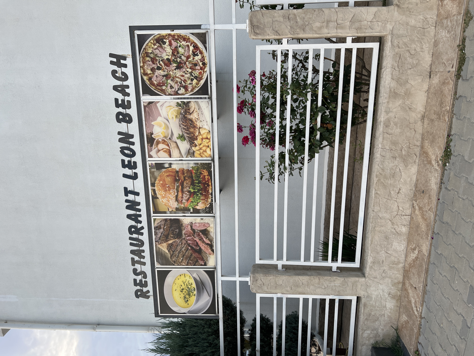 The image shows the exterior of "RESTAURANT LEON BEACH" in Năvodari, Romania. The restaurant's name is displayed in dark, raised lettering on the white exterior wall of a building. Below the name, there are five framed pictures of food items: a bowl of yellow soup, sliced grilled steak, a large hamburger, a grilled fish with lemon slices and bread, and a pizza. The restaurant appears to be located near a street, indicated by the paved sidewalk in the foreground. A white metal fence runs across the middle of the image, separating the building from the street. Behind the fence, there are bushes and some pink flowers. The overall impression is of a casual dining establishment by the sea, suggested by the name "Leon Beach."