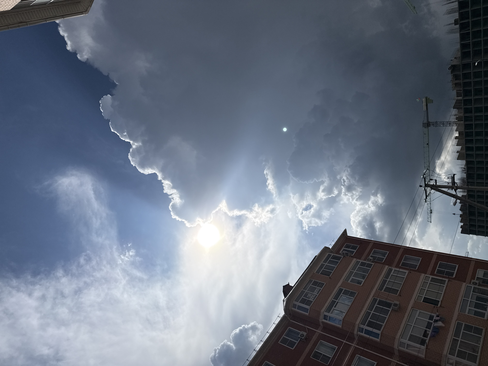 Here is a description of the image:
The image shows a low-angle view of a partially cloudy sky with buildings and a construction crane visible. The sun is obscured by clouds, but its light is still visible, creating a bright spot in the sky. The clouds range from bright white to dark gray, suggesting varying degrees of thickness and density.
A brick building occupies the lower right portion of the frame, with multiple windows visible. To the right and above the brick building, there is a construction site with a partially constructed high-rise building and a construction crane. The environment appears to be urban.