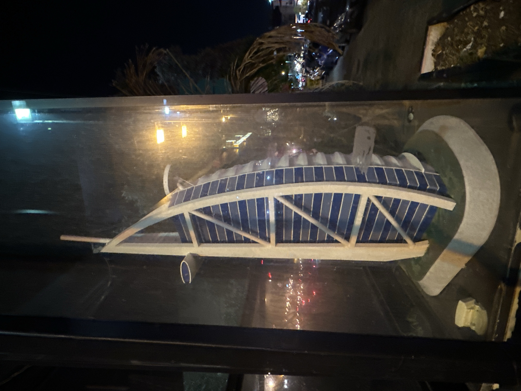 This media file captures a close-up, horizontally oriented, nighttime view of a model of the Burj Al Arab hotel, encased in a glass display. The model is the primary subject, showcasing its distinctive sail-like architecture with blue rectangular panels and white structural supports. The model is positioned on a gray base that mimics the surrounding landscape.
The setting is dark, indicating it is nighttime. The reflection on the glass reveals a blurred background with illuminated lights, suggesting a street or urban environment. The reflections also show other elements of the surrounding environment, such as palm tree fronds and possibly other debris or structures, implying an outdoor or semi-outdoor display location. The lighting within the display is a mix of cooler, brighter lights reflecting from the top and warmer, yellow lights from the sides, highlighting the model.
There are no people visible in the media. The activity taking place is simply the display of the model. Notable details include the reflective nature of the glass enclosure, which adds depth and complexity to the image by overlaying reflections of the surroundings onto the model. The time of day is clearly night, as evidenced by the dark sky and the artificial lighting. No text is visible in the media file. The context provided indicates the location is Haifa, Israel.