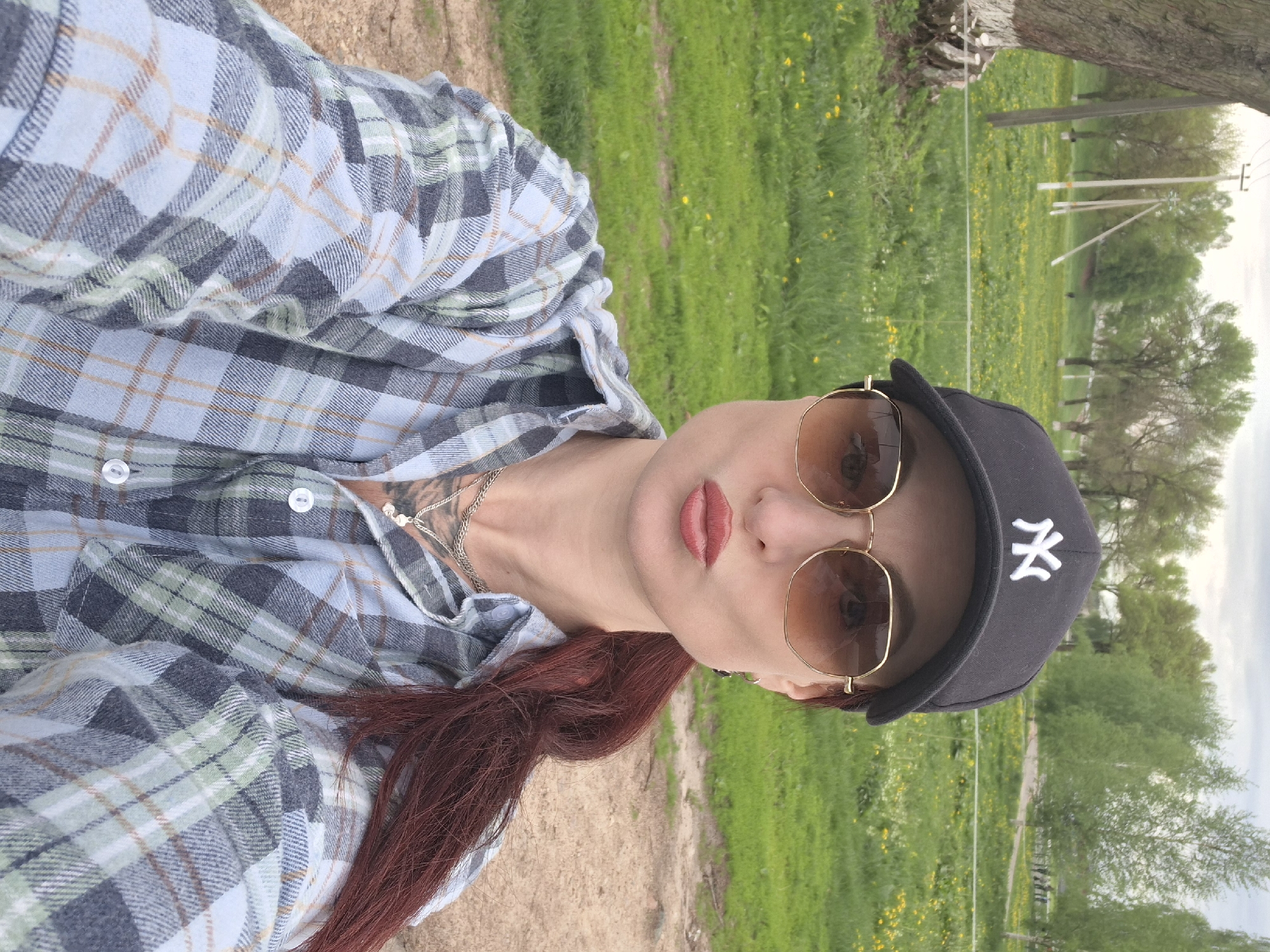 A woman is posing for a selfie outdoors in Забалоцкі сельскі Савет, Belarus. She is wearing a plaid shirt, sunglasses, and a baseball cap with the NY logo. She has long red hair and is wearing red lipstick. The background consists of a grassy field with trees and a dirt path. In the distance, there is a fence and some power lines.