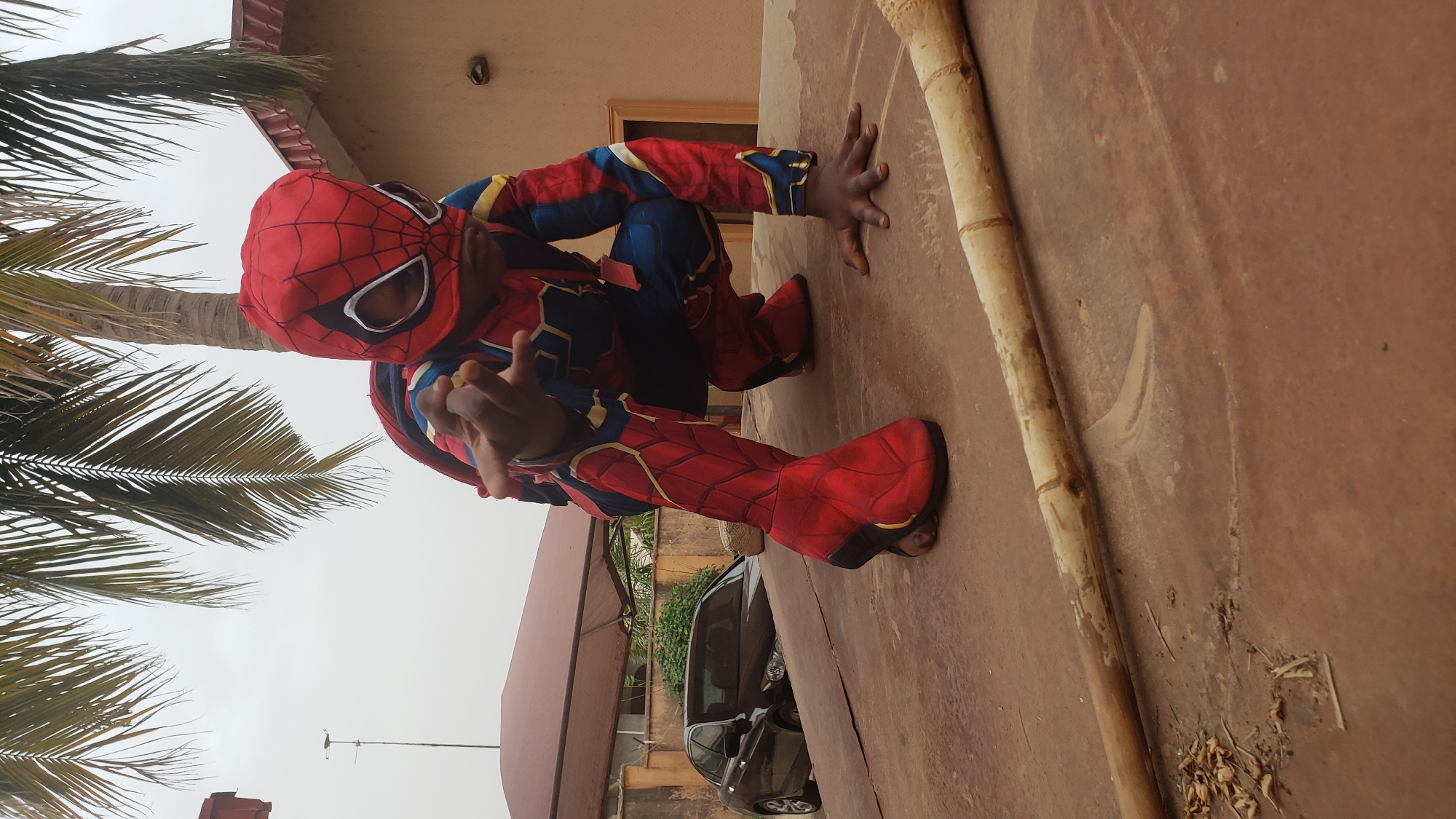 A child in a Spiderman costume is posed crouching on a concrete surface. The person is wearing a red and blue Spiderman suit with a red mask. Their right hand is extended toward the camera, while their left hand is placed on the surface. A light-colored bamboo pole lies next to them on the concrete. Behind them is a tree, a building with a red roof, and a black car. The location is outdoors in Sabon Tasha, Nigeria. The sky appears overcast.