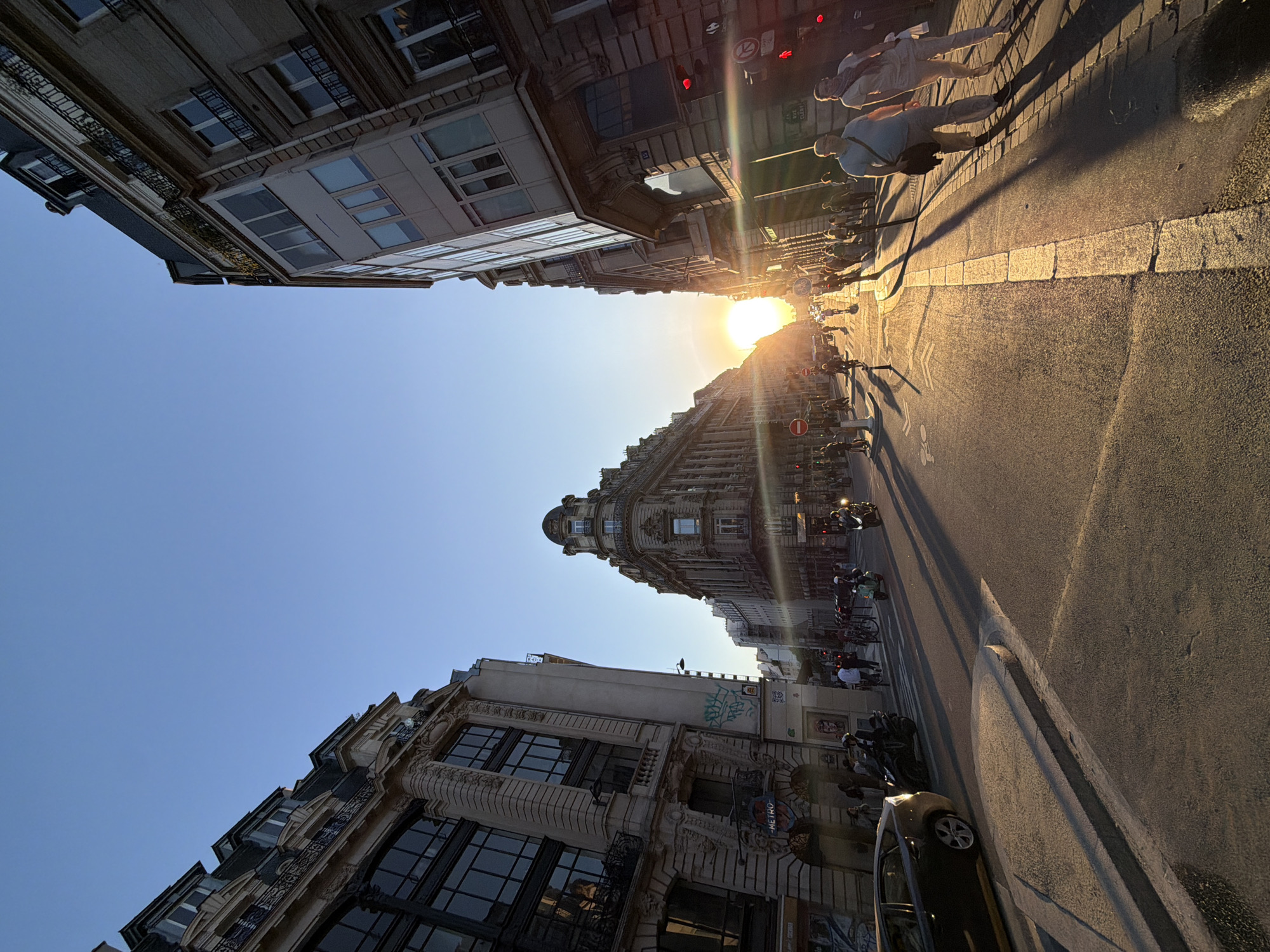 This image captures a vibrant city street scene in Paris, France, during the golden hour, likely sunset, judging by the warm, directional light and long shadows. The sun, a bright orb, is low in the sky, causing a strong lens flare and bathing the street and buildings in a rich golden hue.
The scene features classic Parisian architecture, with multi-story buildings lining both sides of the street. Ornate details, balconies with railings, and large windows are visible. A particularly distinctive building with a rounded, turret-like corner stands out prominently towards the center of the image. The sky above is clear and blue.
On the paved street, traffic appears light, with a few parked cars and numerous scooters or motorcycles visible along the curbs. The asphalt road features white lane markings, including a symbol for a bicycle lane. Pedestrians are scattered on the sidewalks, some silhouetted against the bright light, while others, like a man in a light blue shirt, are illuminated by the low sun, casting long, dramatic shadows behind them.
Notable details include street lights, red traffic lights, and various street signs, such as a "no entry" sign (red circle with white bar) and a "no right turn" sign (red circle with a white arrow). A prominent blue and red circular "METRO" sign, indicating an underground station entrance, is visible on one of the buildings, confirming the urban setting. Some graffiti can be seen on a building wall. The overall atmosphere is warm and active, typical of a bustling city street at the end of the day.