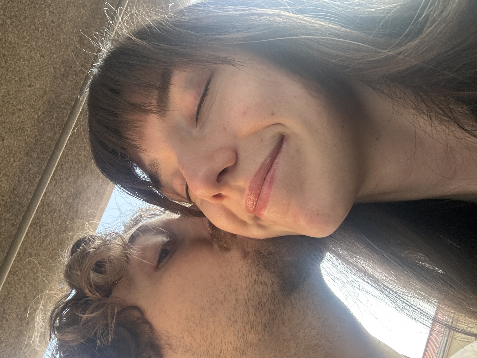 A close-up, selfie-style shot features a couple in a tender moment. The man, on the left, is seen with his eyes closed, leaning in to kiss the woman on the cheek. He has curly brown hair and stubble. The woman, on the right, has her eyes closed and a slight smile, her head tilted slightly upwards. She has brown hair with bangs. The background is an out-of-focus, textured ceiling, possibly from a vehicle or an overhang, with a bright sky visible in the distance. No specific location in Belgrade, Serbia, can be identified from the image.