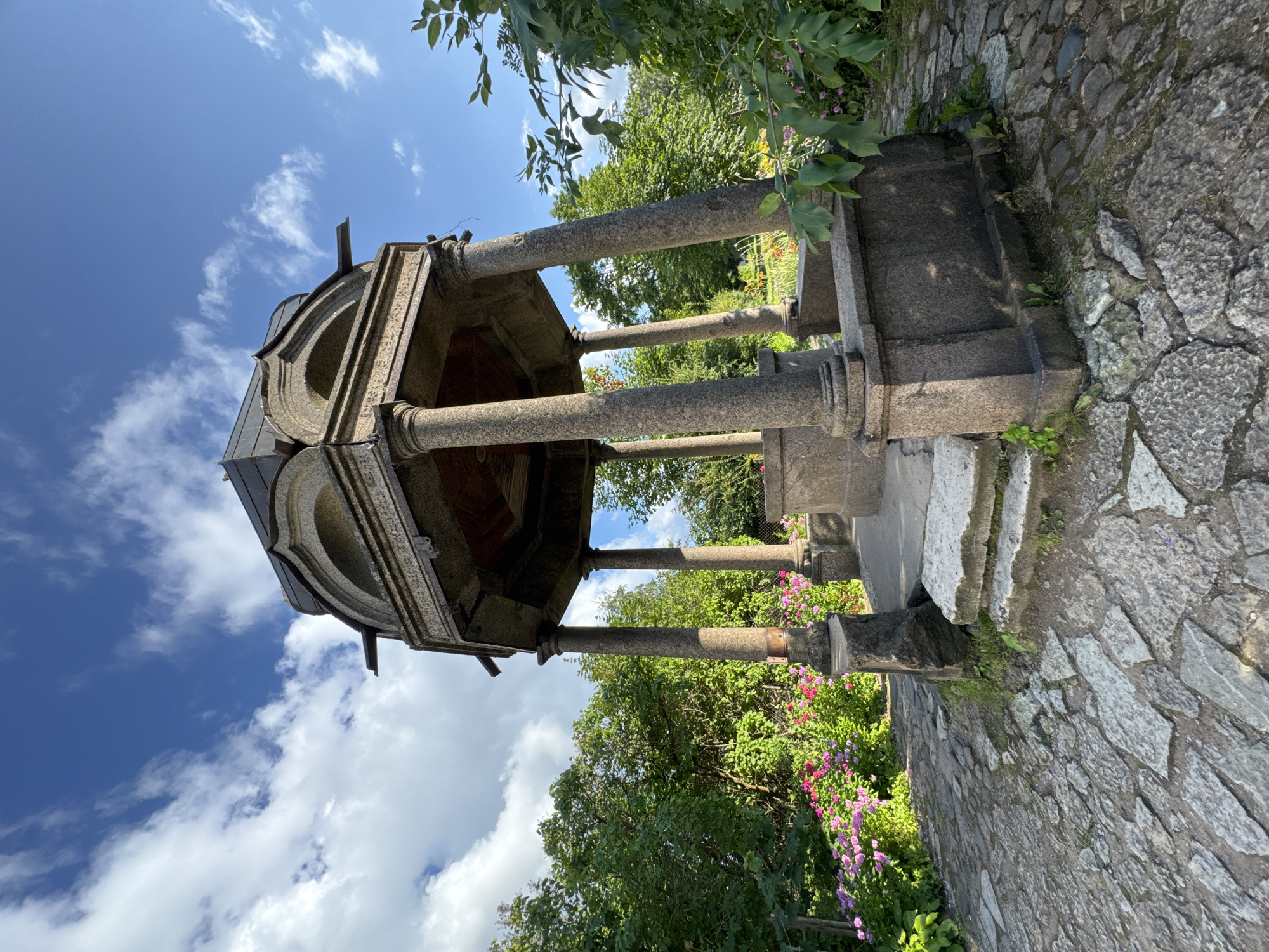 The image shows an octagonal stone pavilion with a domed roof and four stone columns supporting it. The pavilion is situated in a garden setting with lush green trees and flowering bushes in the background. The ground is paved with irregular stones. The sky above is blue with scattered white clouds. The pavilion appears to be an old structure, possibly a historical monument. Based on the architecture and garden style, this could be a gazebo or a folly found in a park or estate. Without further information, the specific location in Veliky Novgorod, Russia, cannot be definitively identified from the image alone.