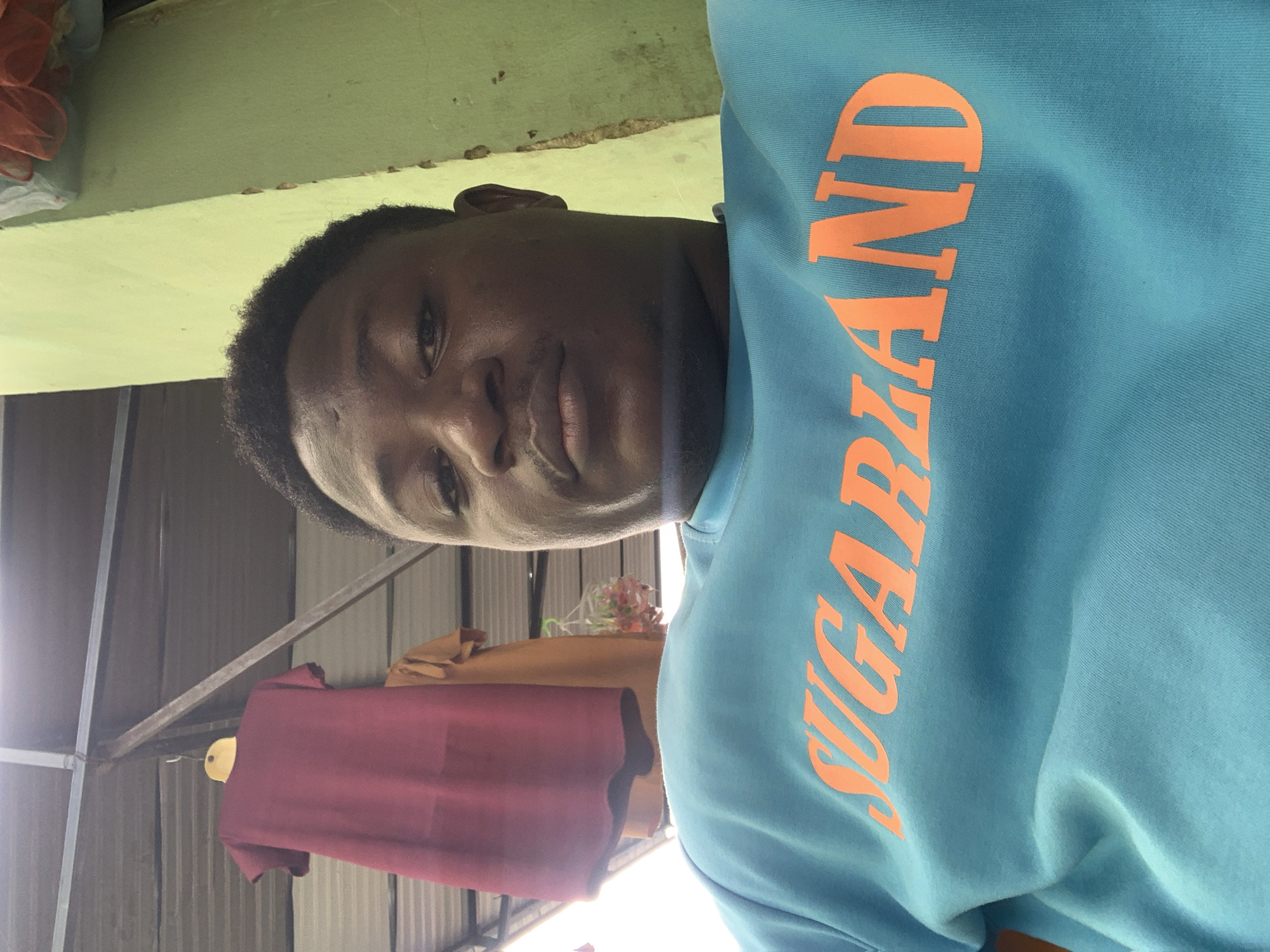 In a Nigerian outdoor setting, a close-up shot focuses on a person wearing a light blue sweatshirt with "SUGARLAND" emblazoned in orange lettering. The background reveals a light green wall and a corrugated metal roof supported by metal beams. A maroon shirt hangs on a yellow hanger in the upper left corner. The image appears to be taken in Akwanga, Nigeria.