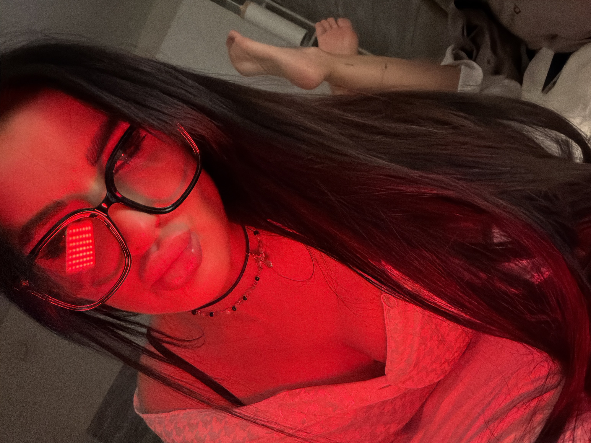 A person is lying down, facing the camera. The person has long, dark hair and is wearing glasses with a red, grid-like pattern visible in one lens. Their lips are slightly parted, and they are wearing a necklace. The upper part of their body is visible, covered by a light-colored patterned shirt. In the background, slightly out of focus, are feet. The environment is illuminated by red light. There is no information to determine whether this image was taken in Moscow.