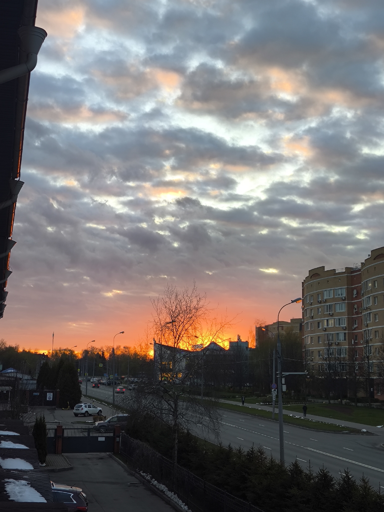 The image presents an urban street scene captured at sunset, likely from an elevated perspective such as a window or balcony in Moscow, Russia.
The most striking subject is the sky, ablaze with the intense, fiery orange and red glow of the setting sun near the horizon. This vibrant light diffuses into softer pinks, blues, and grays within the scattered, overcast cloud formations overhead.
In the immediate foreground on the left, a portion of a dark roof and a white downspout are visible. Below, a gated driveway leads to a multi-lane road. Patches of snow on the ground and rooftops suggest a late winter or early spring setting.
The midground features a busy road with cars, including a prominent white SUV, moving and parked. Streetlights are visible, some already active. Bare trees line the road, interspersed with some evergreen shrubs. On the right, a large, multi-story apartment building with numerous windows and balconies dominates the urban landscape. Two individuals are discernible walking on the sidewalk adjacent to the road.
The activity taking place includes vehicles driving on the road and people walking. The time of day is clearly sunset, casting long shadows and dramatic light across the scene. The architecture of the apartment building and the general layout of the wide road are consistent with urban environments in Moscow. There is no visible text in the image.