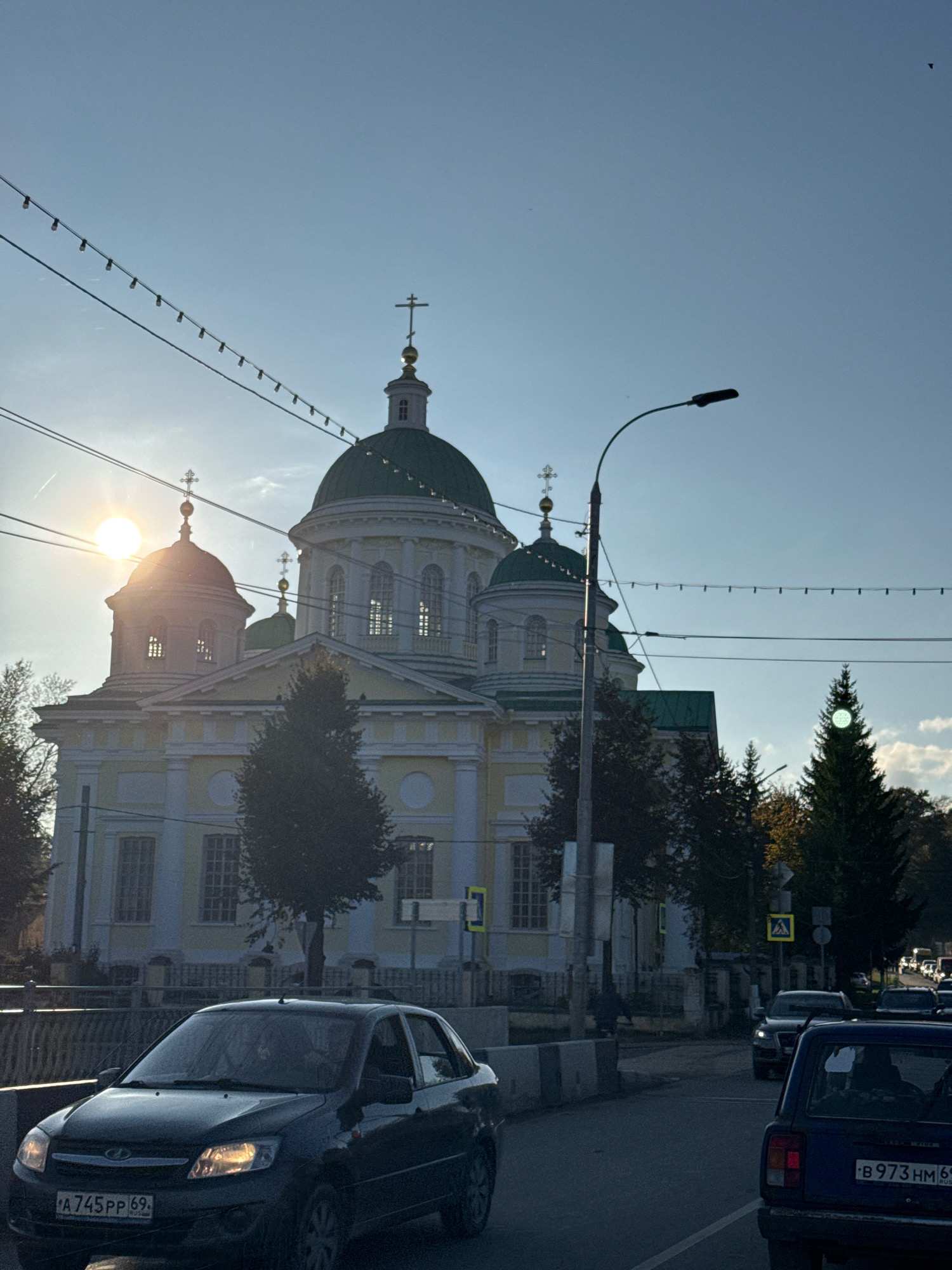 The image depicts a view of the Borisoglebsky Monastery in Torzhok, Russia, under a clear blue sky with the sun low on the horizon, creating a lens flare. The monastery features several domes, one prominent green dome with a cross, and lighter colored walls with arched windows. In the foreground, a road runs in front of the monastery, with cars visible. A dark sedan with license plate "A 745 PP 69" is in the left lane, and a blue car with license plate "B 973 HM 69" is in the right lane. Power lines with decorative lights are strung across the sky. Trees line the side of the road and are visible around the monastery. The overall scene suggests a late afternoon or early evening setting.